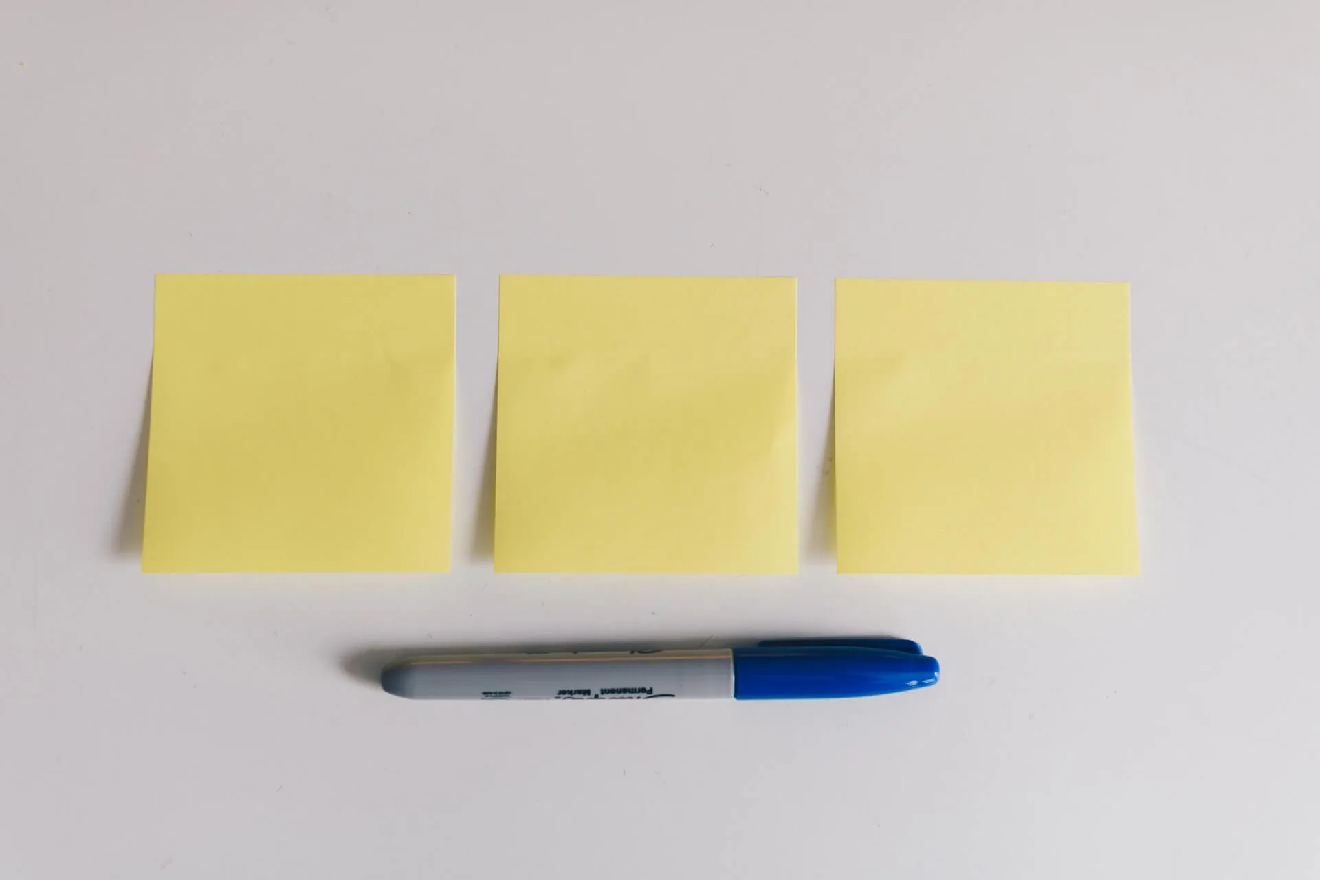 Three blank yellow sticky notes aligned side by side above a grey permanent marker with a blue cap on a white surface