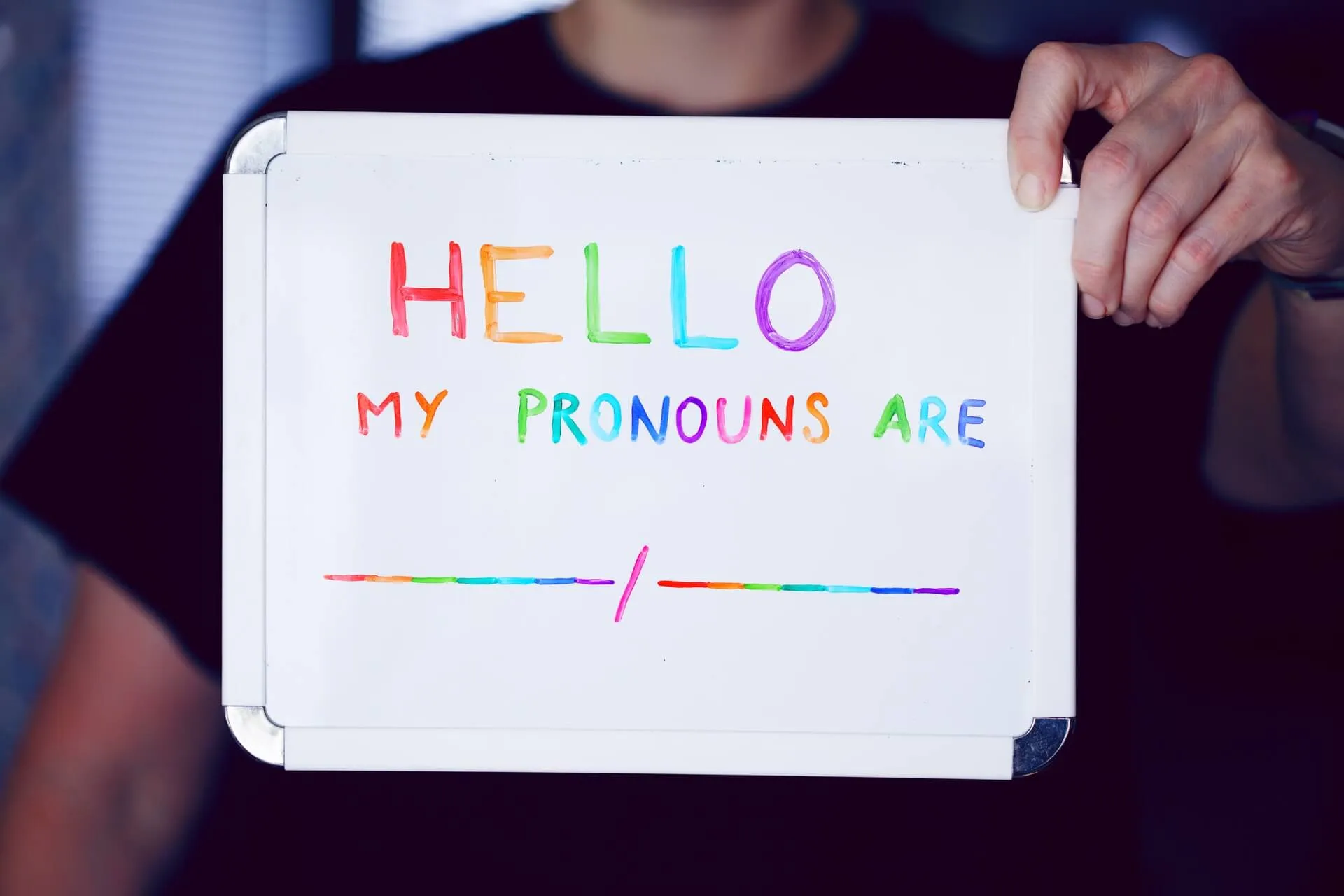 Hands holding a whiteboard with hand drawn rainbow letters spelling HELLO my pronouns are followed by two blank rainbow lines