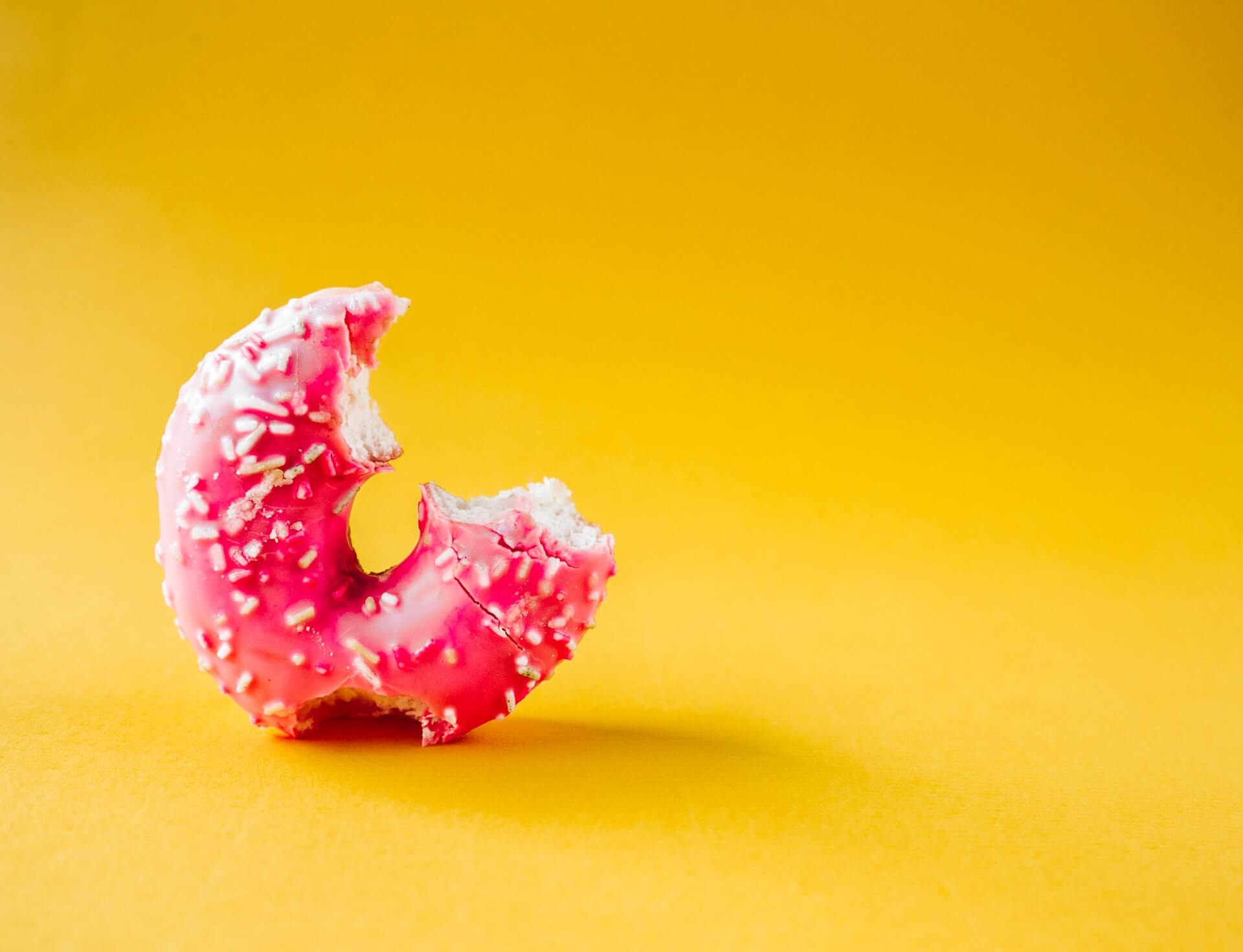 Close-up of a pink-glazed donut topped with white sprinkles and one large bite taken, set against a solid yellow backdrop
