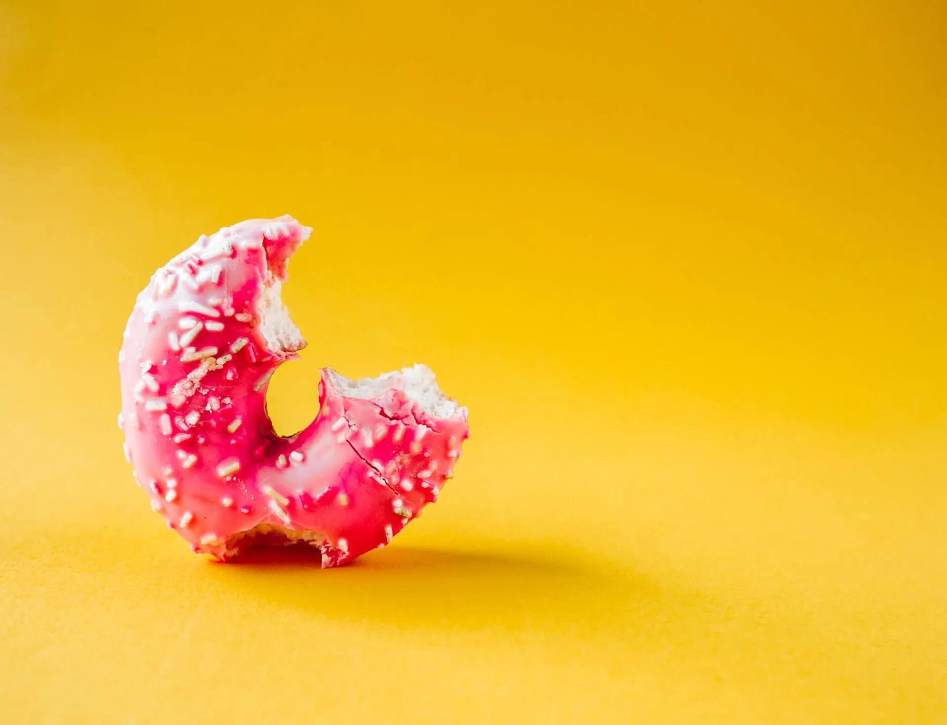 Close-up of a pink-glazed donut topped with white sprinkles and one large bite taken, set against a solid yellow backdrop
