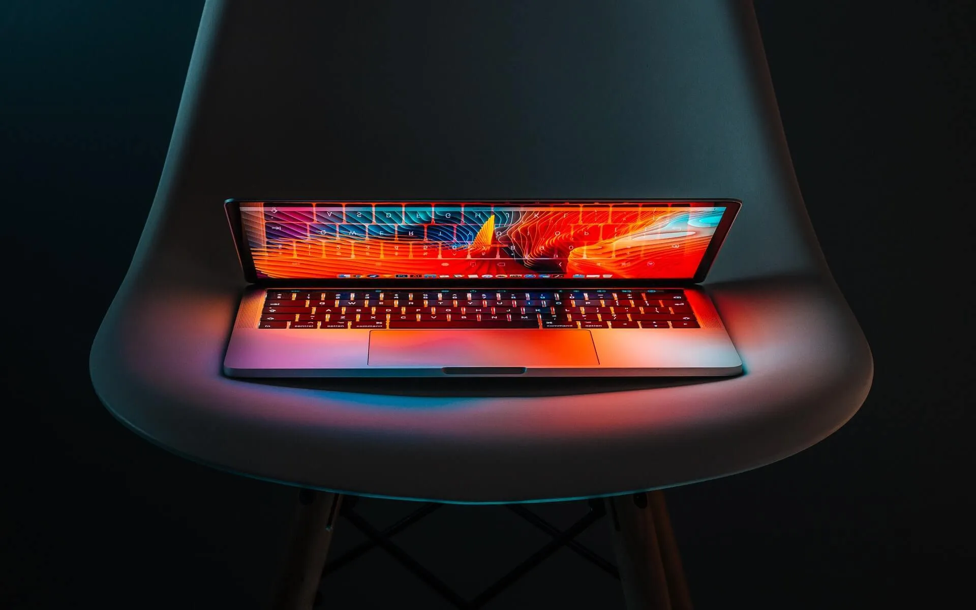 Partially open laptop resting on a curved white chair bathed in striking red and blue light reflecting across its keyboard and seat