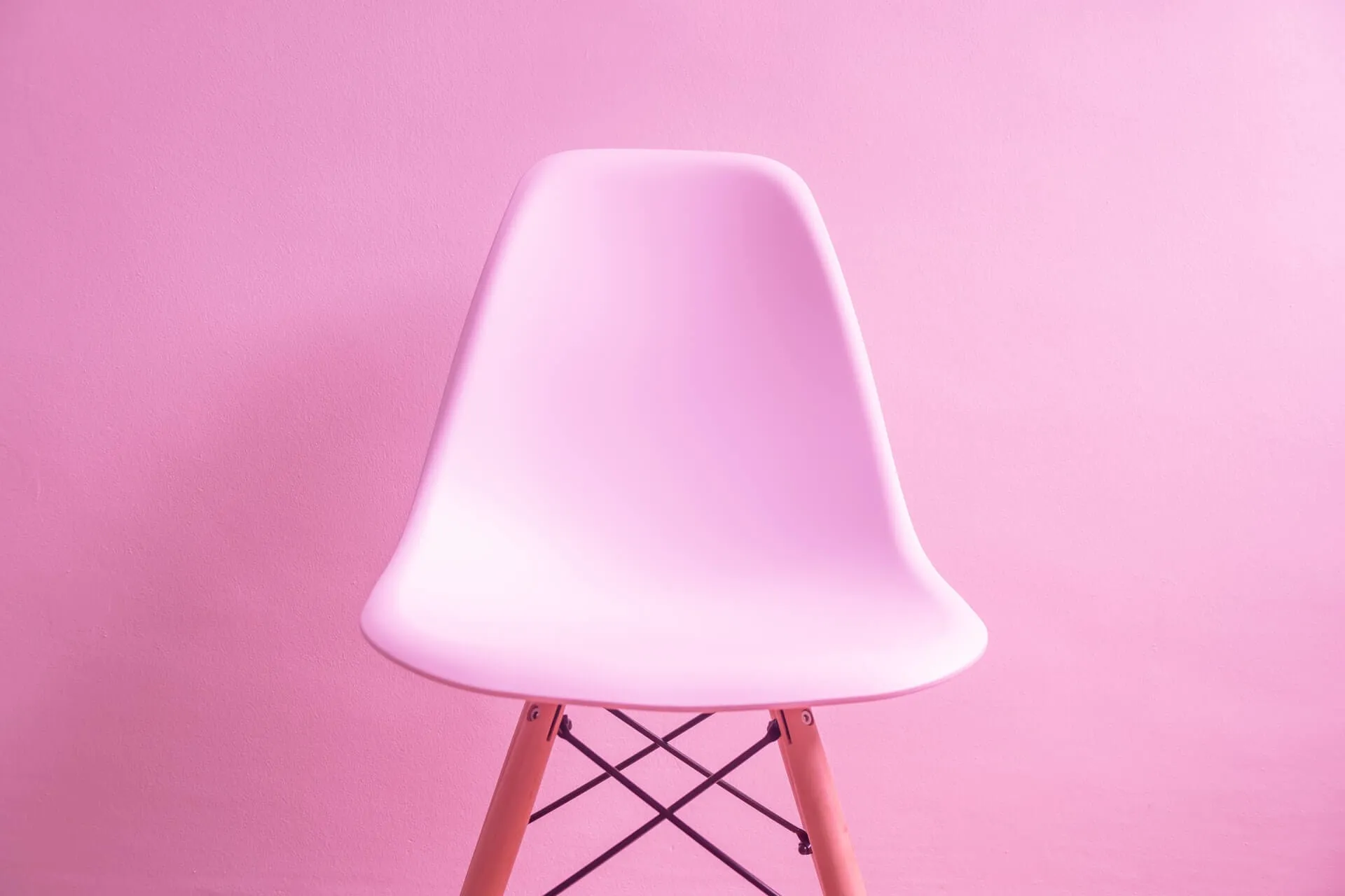 Single pastel pink plastic chair featuring wooden dowel legs and metal supports positioned against a matching pastel pink background