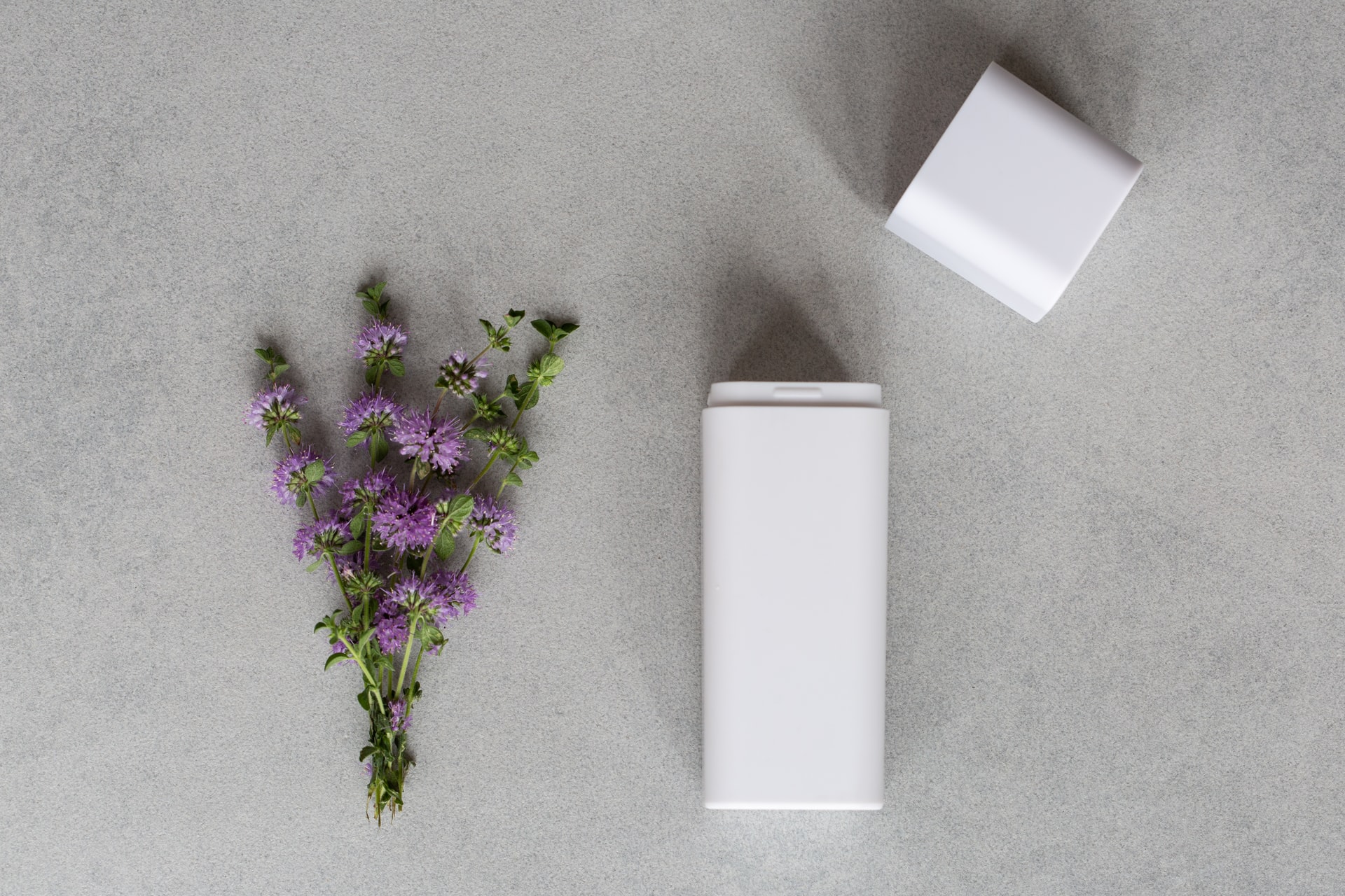Overhead view of a white rectangular deodorant container with its cap removed placed next to a small bouquet of purple mint flowers on a grey stone surface