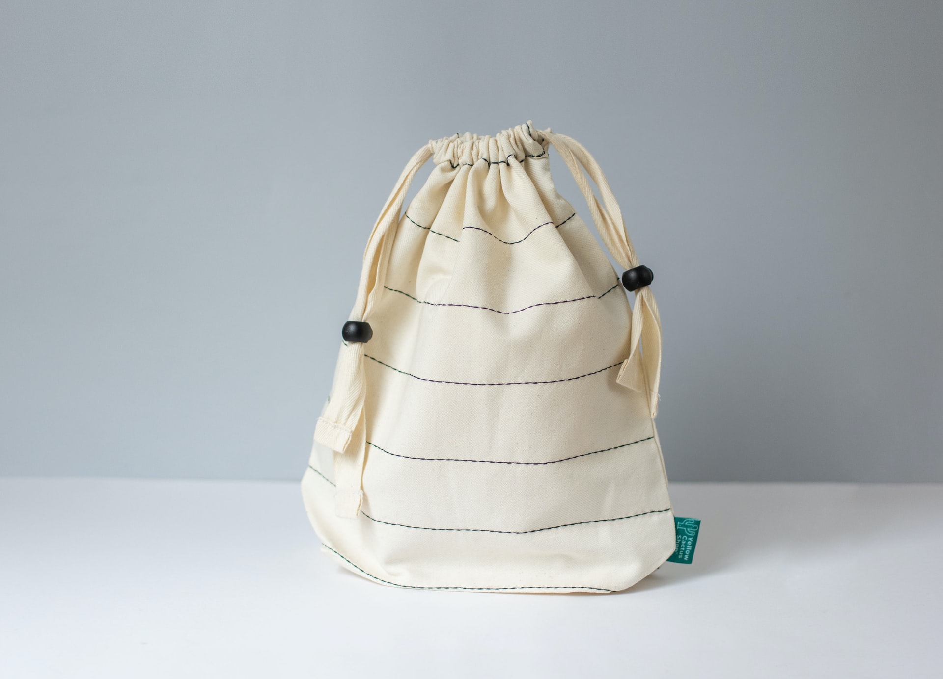 Cream canvas drawstring pouch bag with subtle horizontal stitching lines and black bead cinch toggles on a white tabletop against a gray backdrop