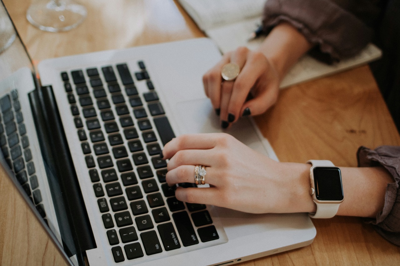 Closeup of hands with dark nail polish rings and smartwatch typing on a silver laptop on a wooden desk with a notebook and glass in view