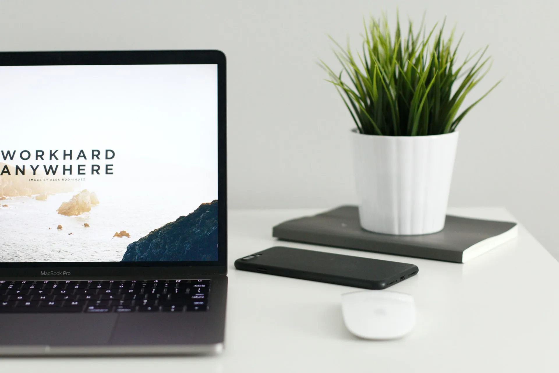MacBook Pro on a white desk displaying a coastal scene with the words work hard anywhere next to an Apple mouse a smartphone a closed notebook and a white potted plant