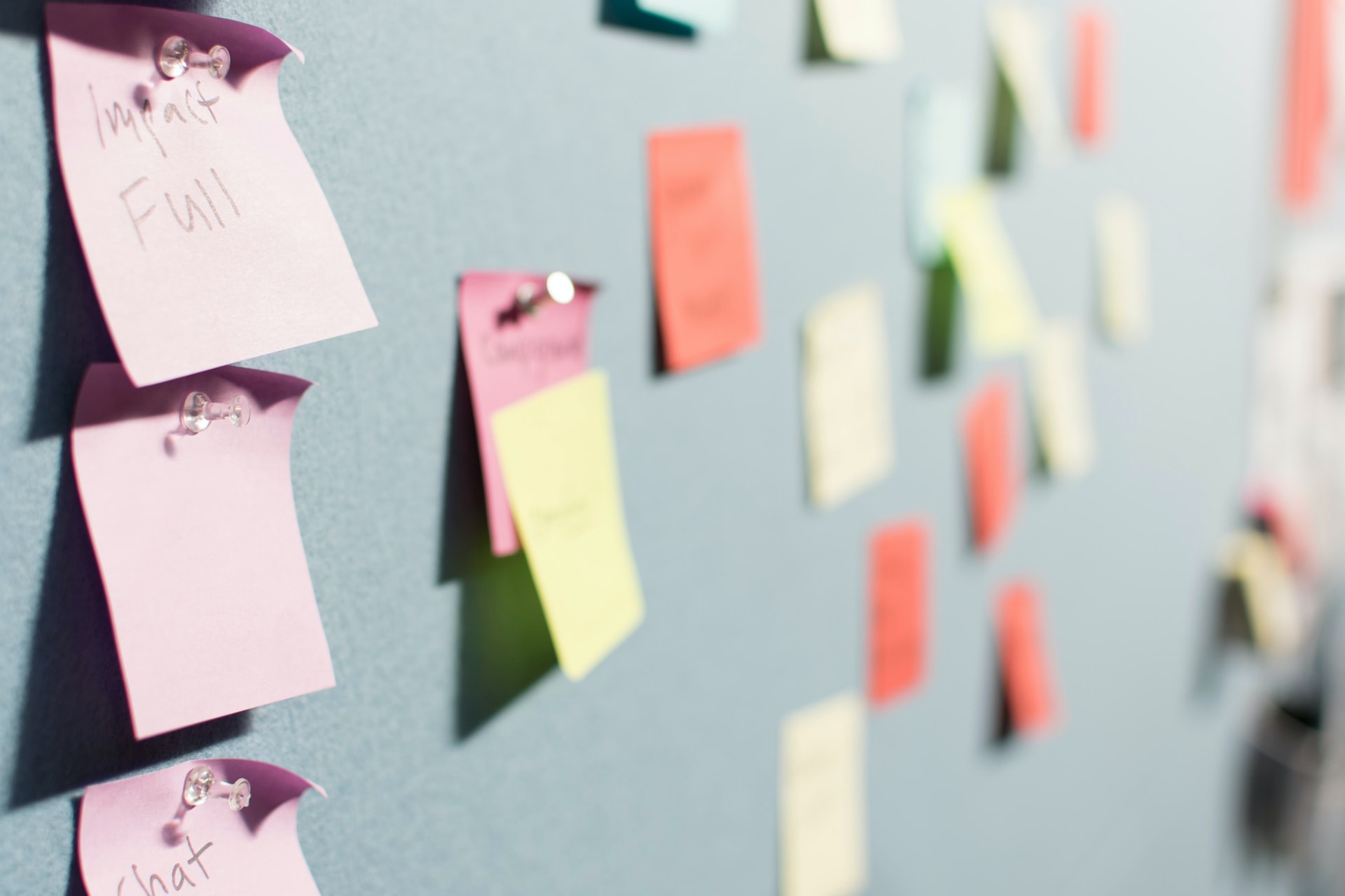 Close up of a gray pin board with pink sticky notes pinned in a column and blurred multi colored notes behind evoking a brainstorming board