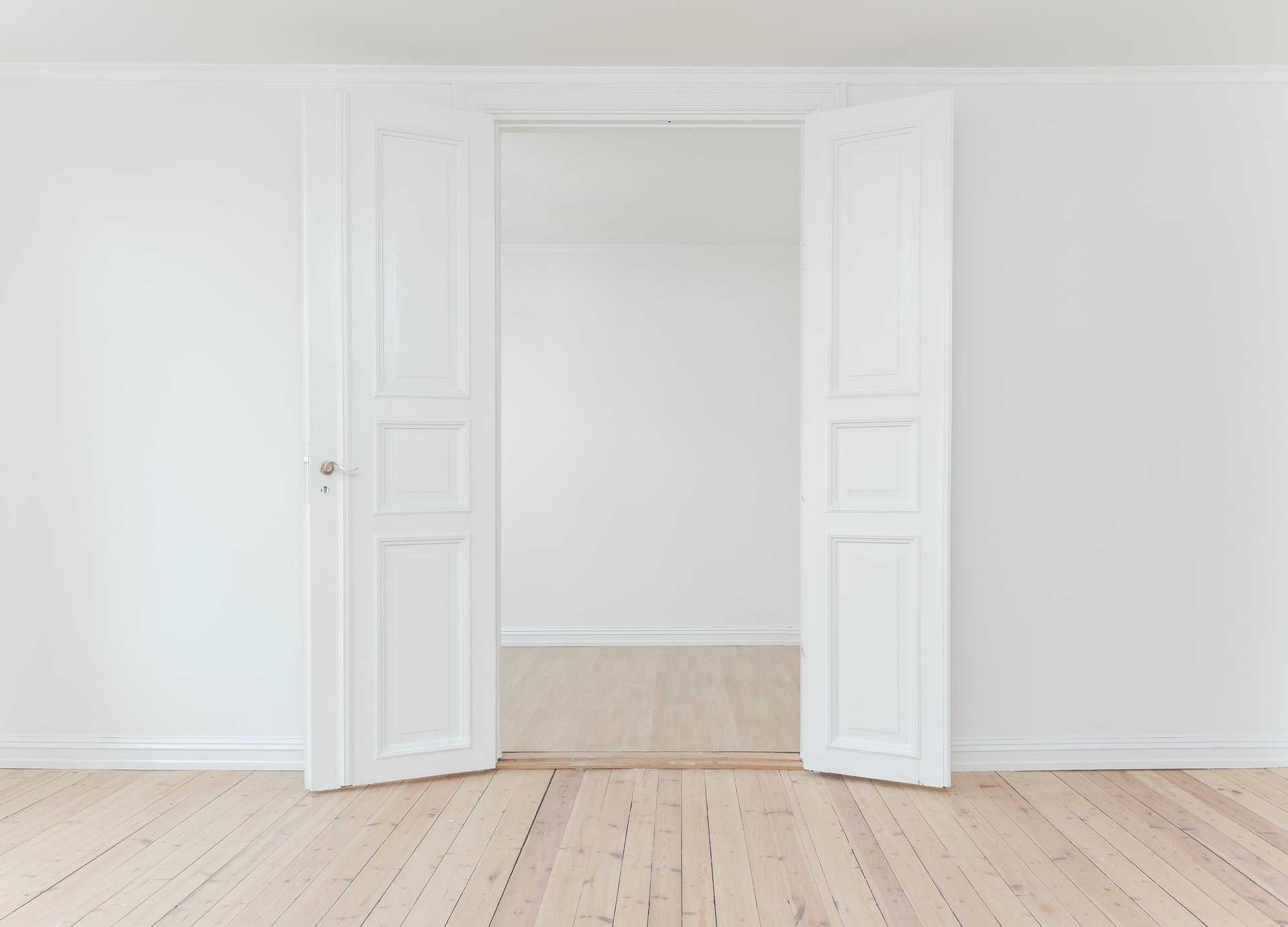 Straight-on view of open ornate white double doors revealing an empty white-walled room with light wooden floors and matching trim, suggesting a bright, minimalist interior space