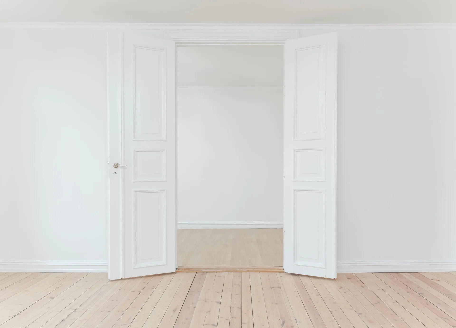 Straight-on view of open ornate white double doors revealing an empty white-walled room with light wooden floors and matching trim, suggesting a bright, minimalist interior space