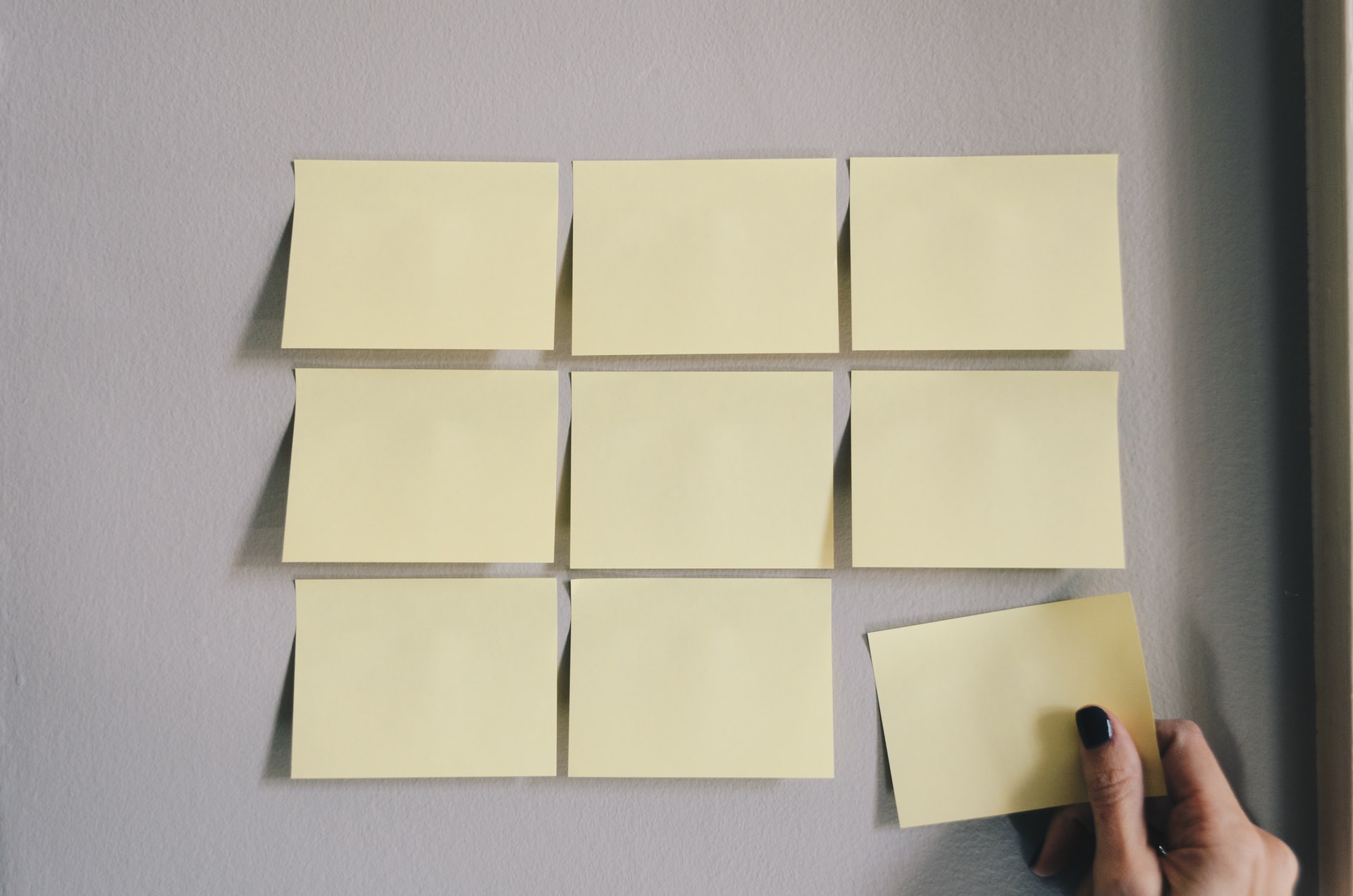 Close-up of blank yellow sticky notes in three rows on a light grey wall, showing a hand placing the bottom-right note