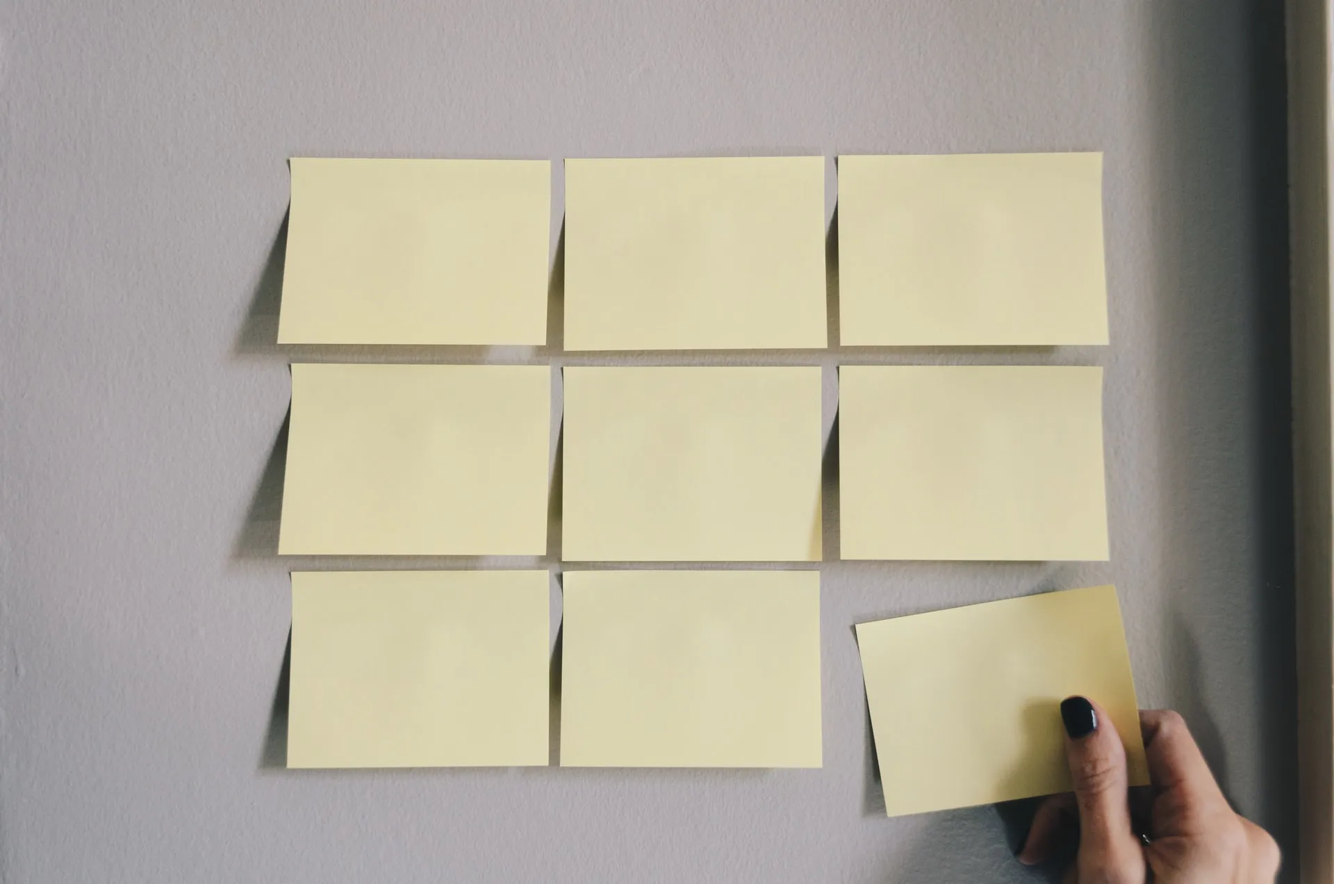 Close-up of blank yellow sticky notes in three rows on a light grey wall, showing a hand placing the bottom-right note