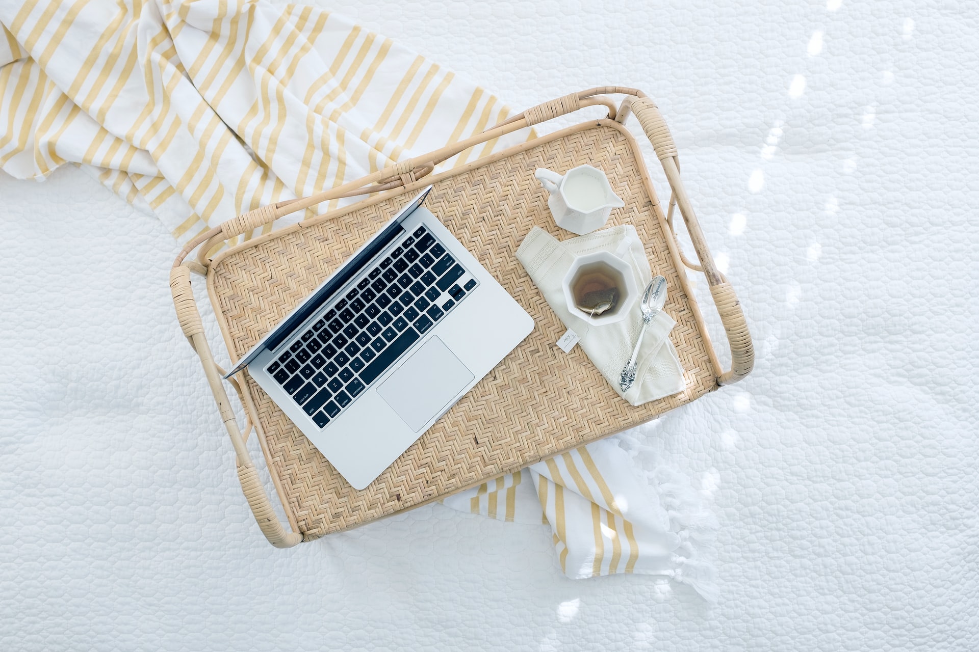 Overhead view of a woven rattan serving tray on a white quilted bed with a yellow striped throw edge, featuring an open laptop, a cup of tea with a spoon and a small white creamer jug