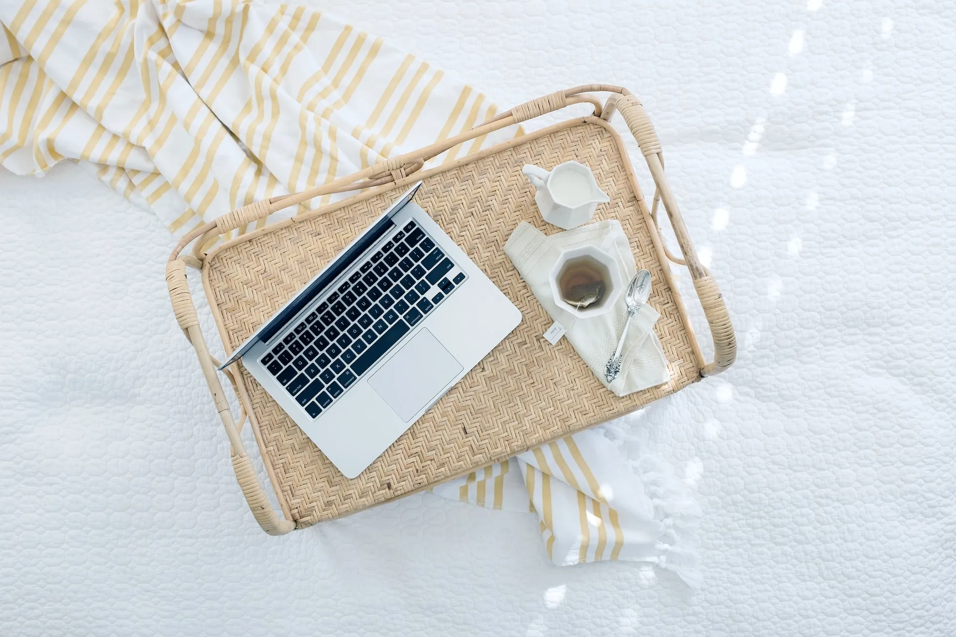 Overhead view of a woven rattan serving tray on a white quilted bed with a yellow striped throw edge, featuring an open laptop, a cup of tea with a spoon and a small white creamer jug