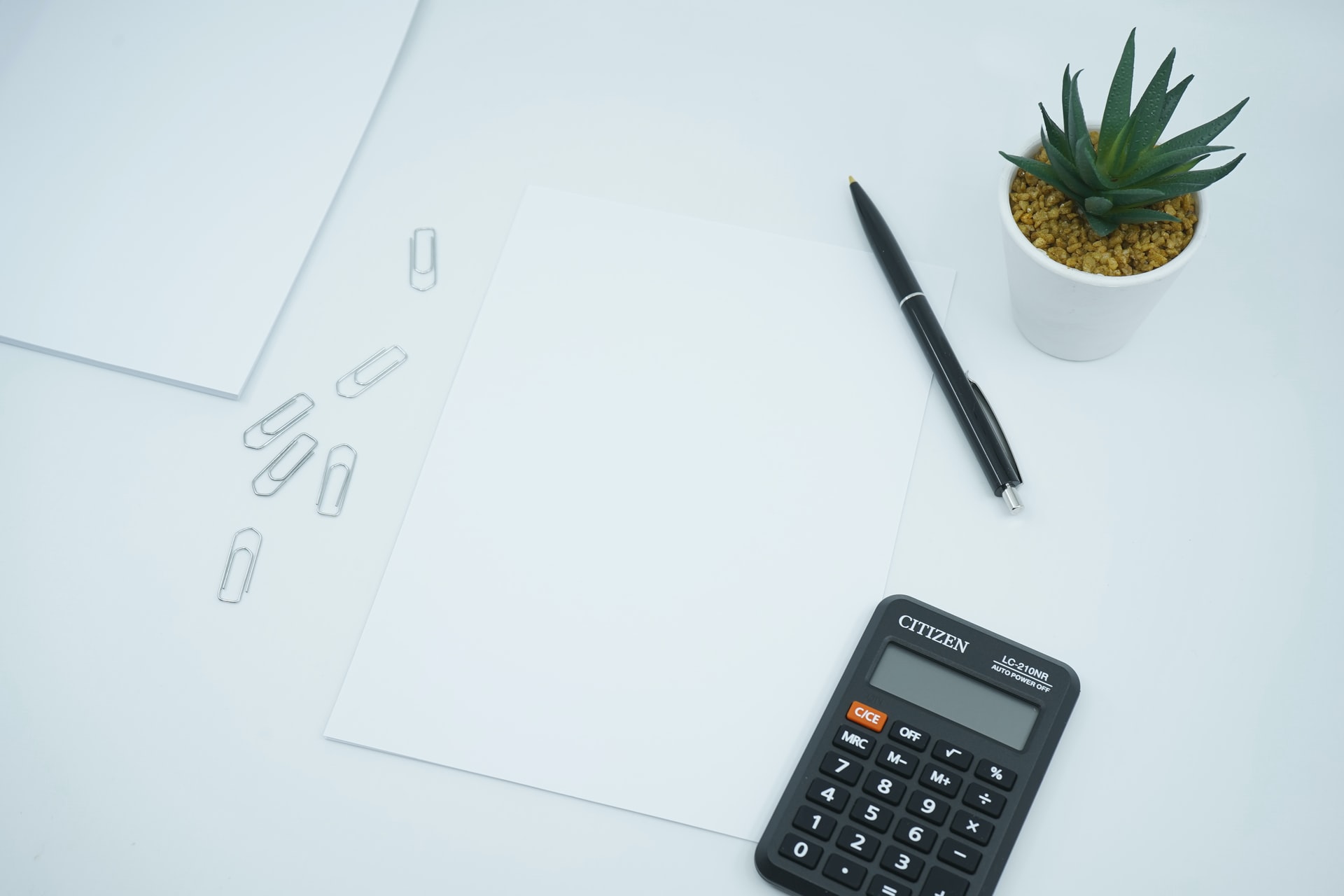 Clean white workspace seen from above, featuring a blank sheet of paper at center, scattered silver paper clips, a black ballpoint pen, a Citizen calculator and a small green succulent in a white pot