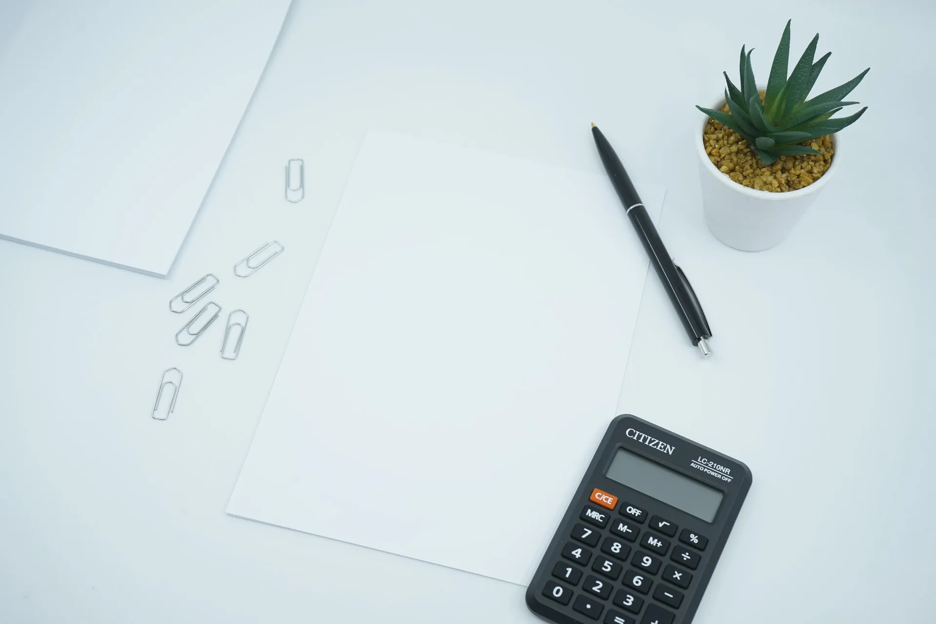 Clean white workspace seen from above, featuring a blank sheet of paper at center, scattered silver paper clips, a black ballpoint pen, a Citizen calculator and a small green succulent in a white pot