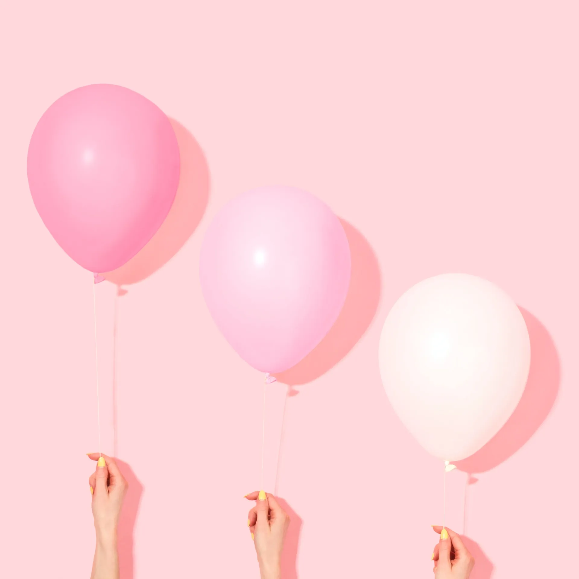 Three balloons in pastel pink, light pink and pale white held by hands with yellow nail polish against a light pink background