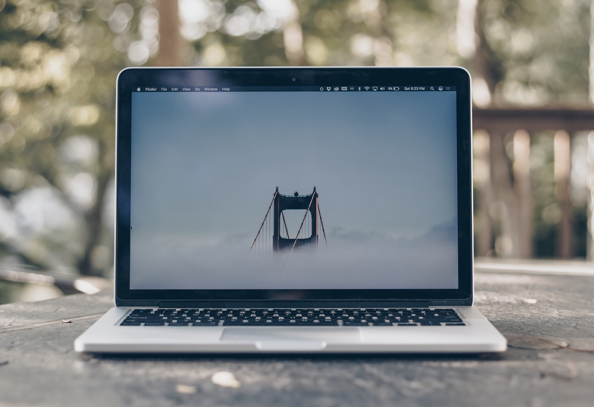 Silver laptop on a wooden outdoor table with blurred green trees in the background displays the Golden Gate Bridge tower rising through fog on its screen