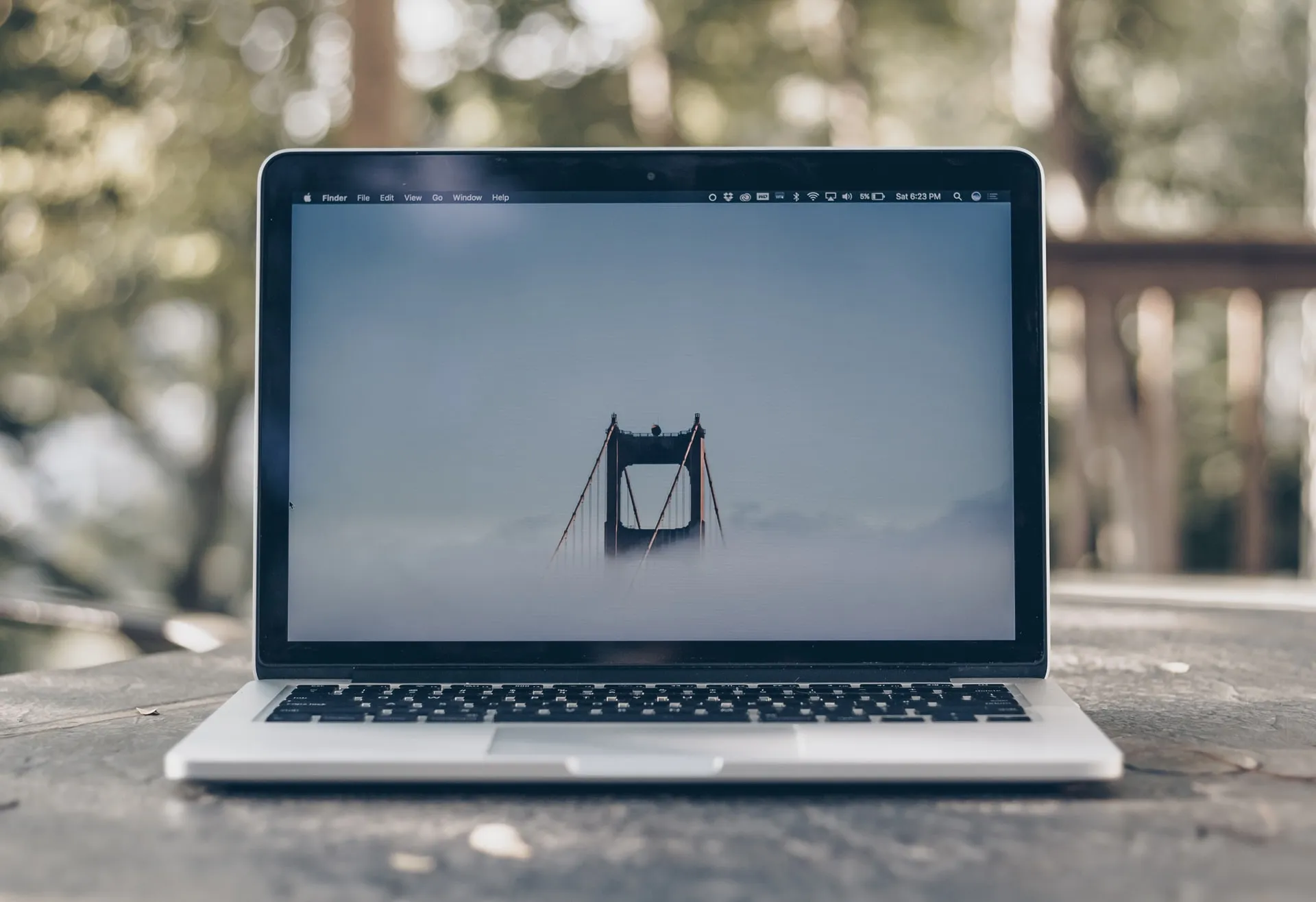 Silver laptop on a wooden outdoor table with blurred green trees in the background displays the Golden Gate Bridge tower rising through fog on its screen