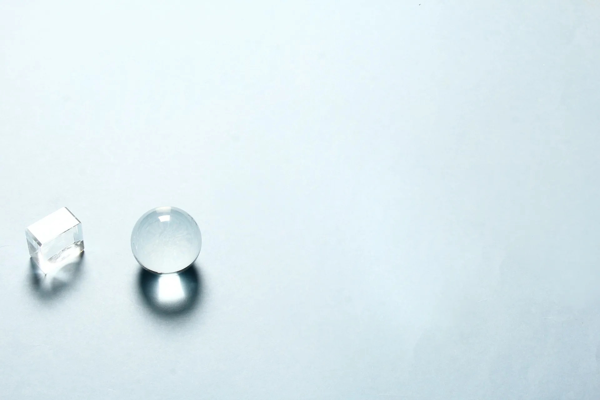 Clear glass sphere next to a rectangular prism casting soft shadows on a light grey background