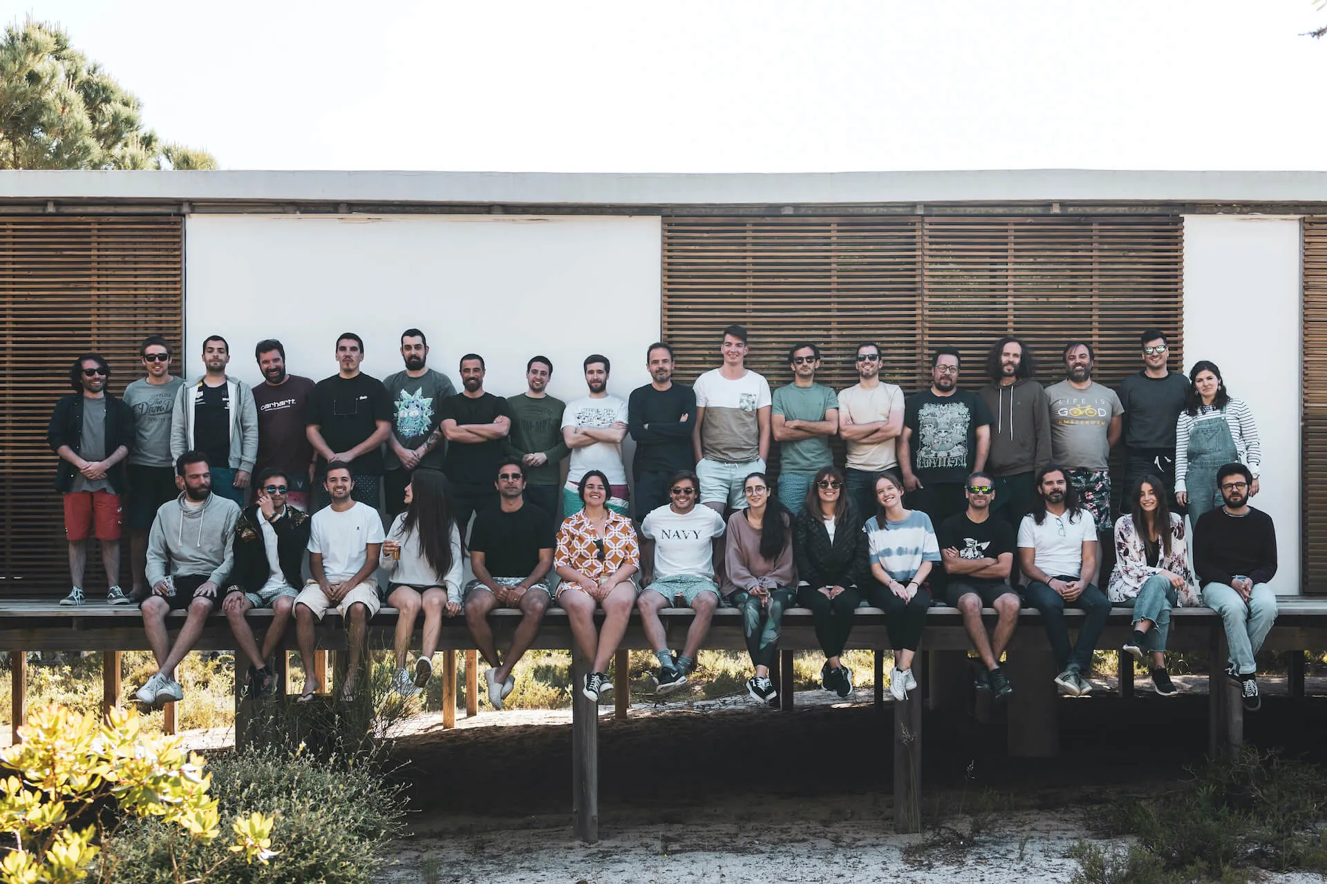 Coverflex team around thirty people arranged in two rows on a raised wooden deck outside a minimalist cabin by the beach smiling and posing with casual summer attire