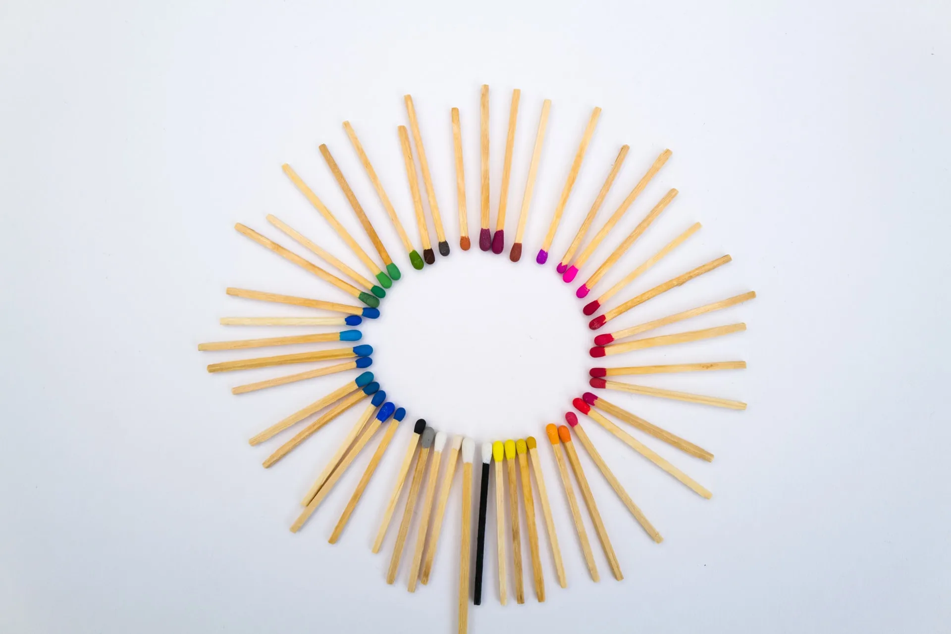 overhead view of a ring of wooden matchsticks with heads in rainbow colors on a white background surrounding one dark burnt matchstick at the bottom center