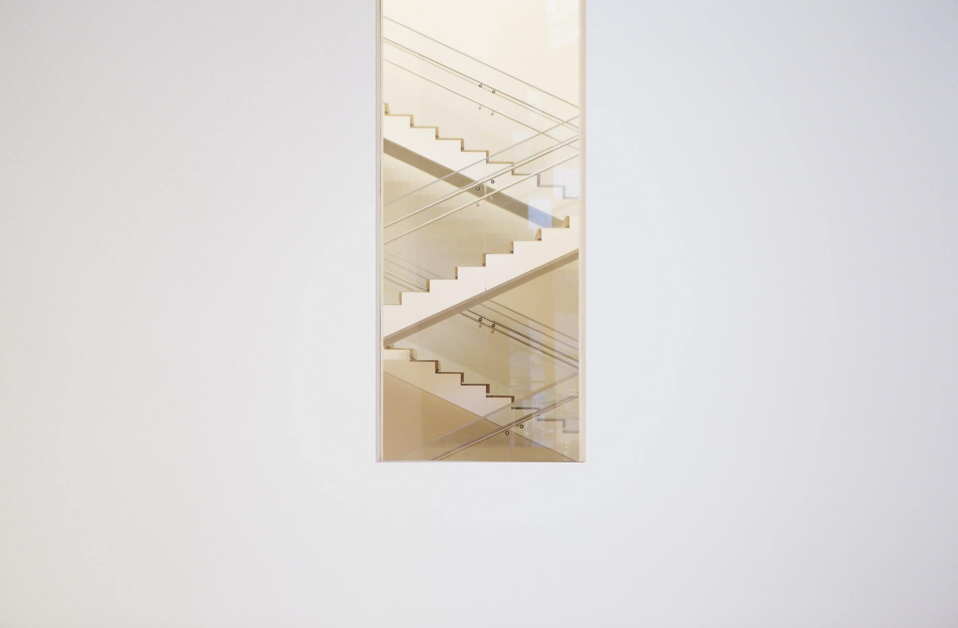 view through a narrow vertical opening in a plain white wall showing intersecting indoor staircases with glass handrails and soft golden light