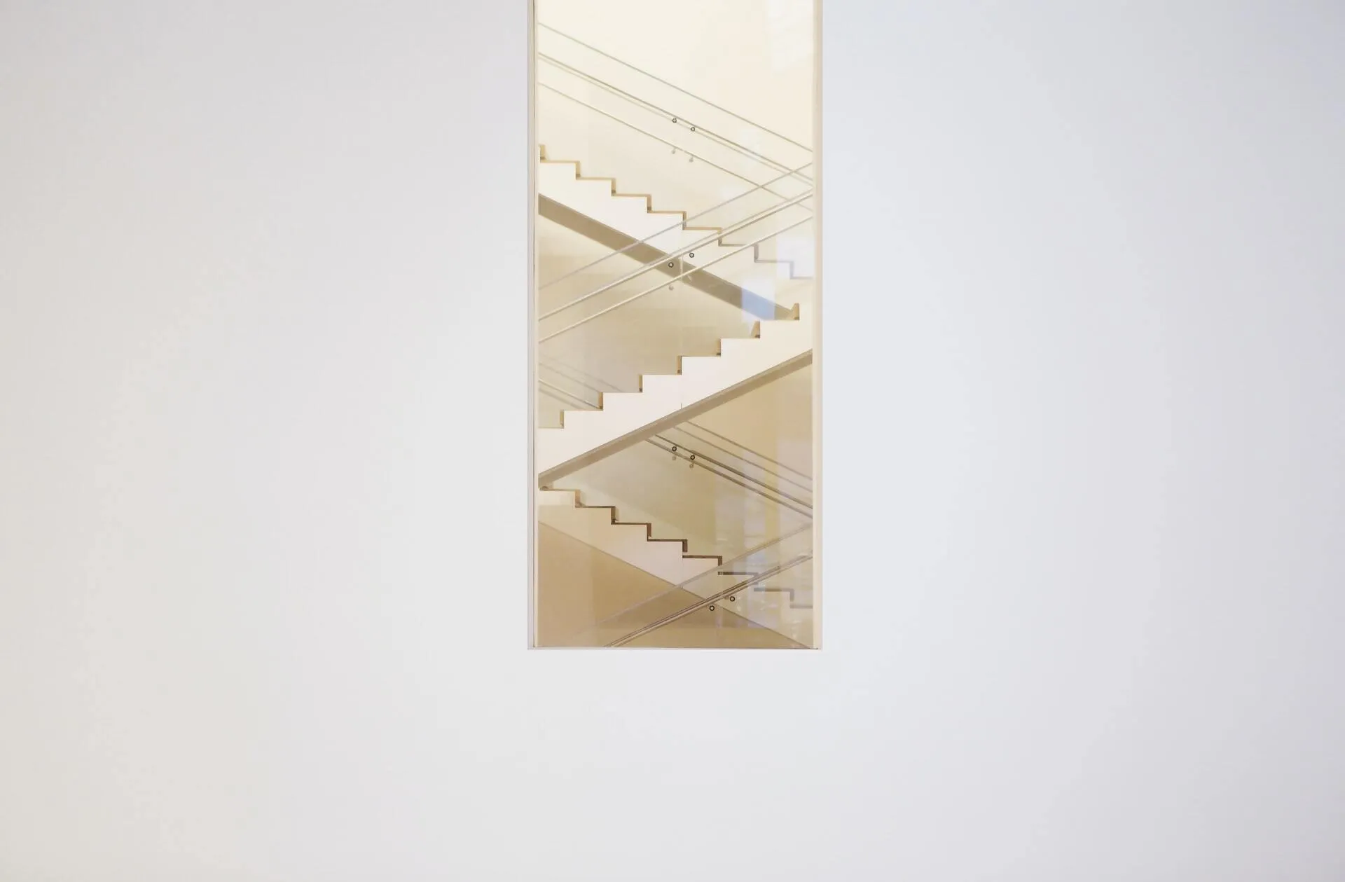 view through a narrow vertical opening in a plain white wall showing intersecting indoor staircases with glass handrails and soft golden light