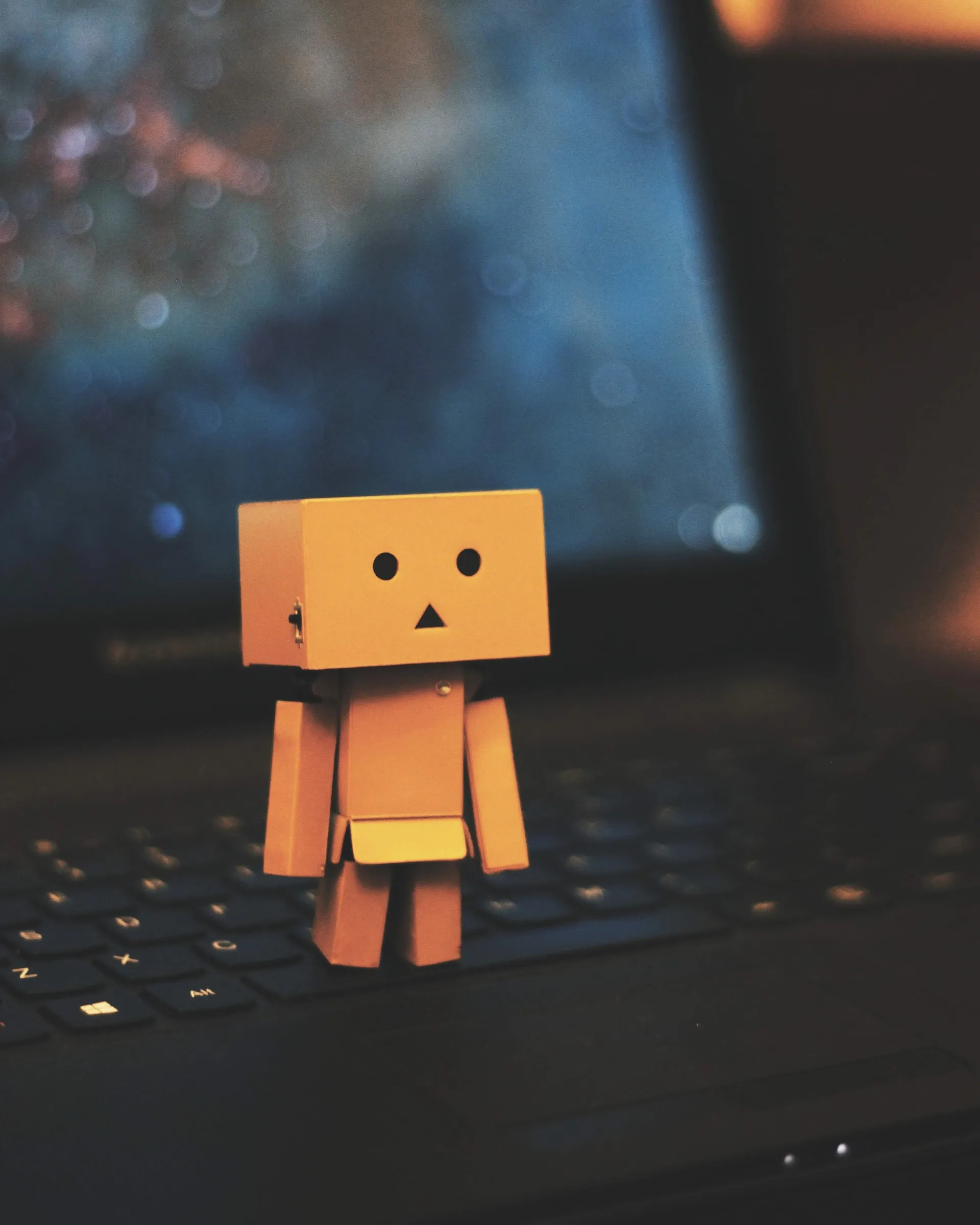 Danbo cardboard toy robot standing on a laptop keyboard in warm ambient light with a blurred screen in the background