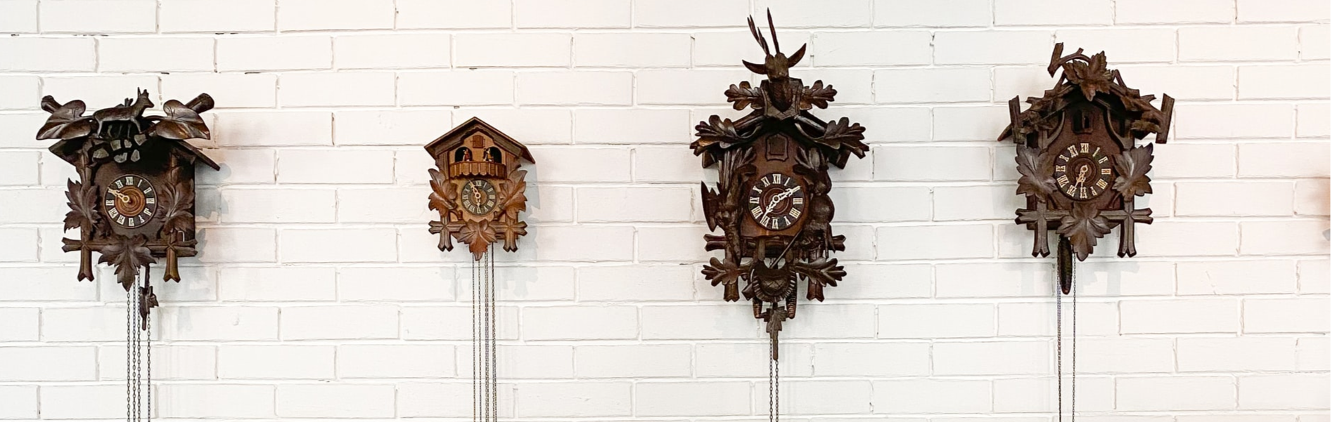 Row of four ornate wooden cuckoo clocks featuring leaf carvings and animal figures mounted on a white painted brick wall