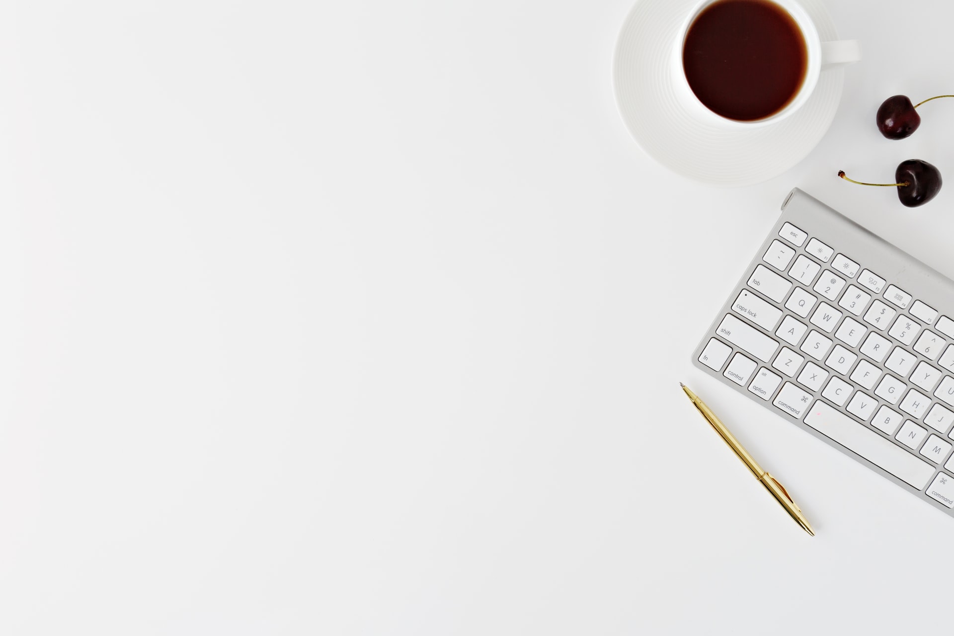 overhead shot showing a white wireless keyboard and gold pen beside a white coffee cup filled with coffee and two dark cherries on a clean white desktop with ample empty space