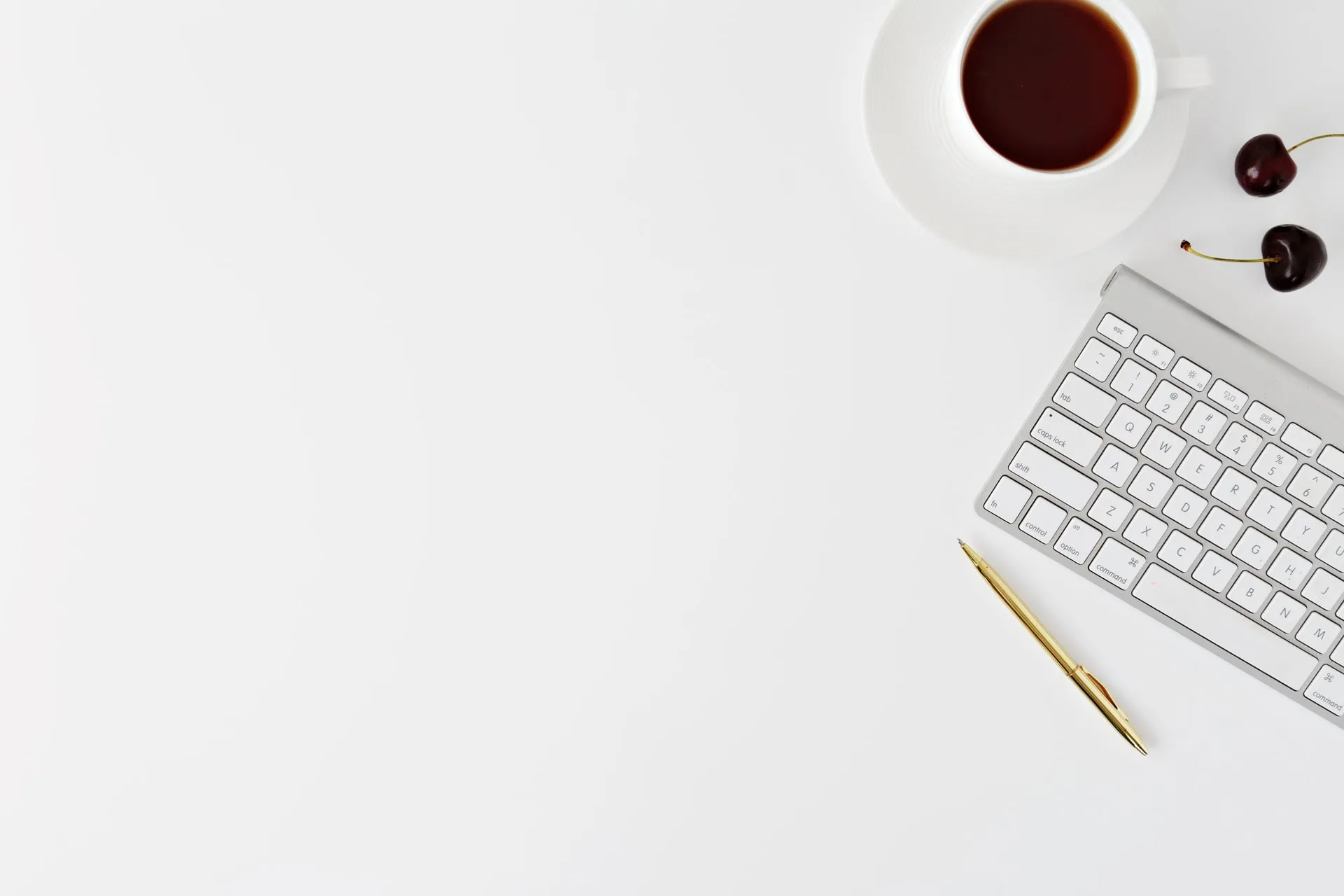 overhead shot showing a white wireless keyboard and gold pen beside a white coffee cup filled with coffee and two dark cherries on a clean white desktop with ample empty space