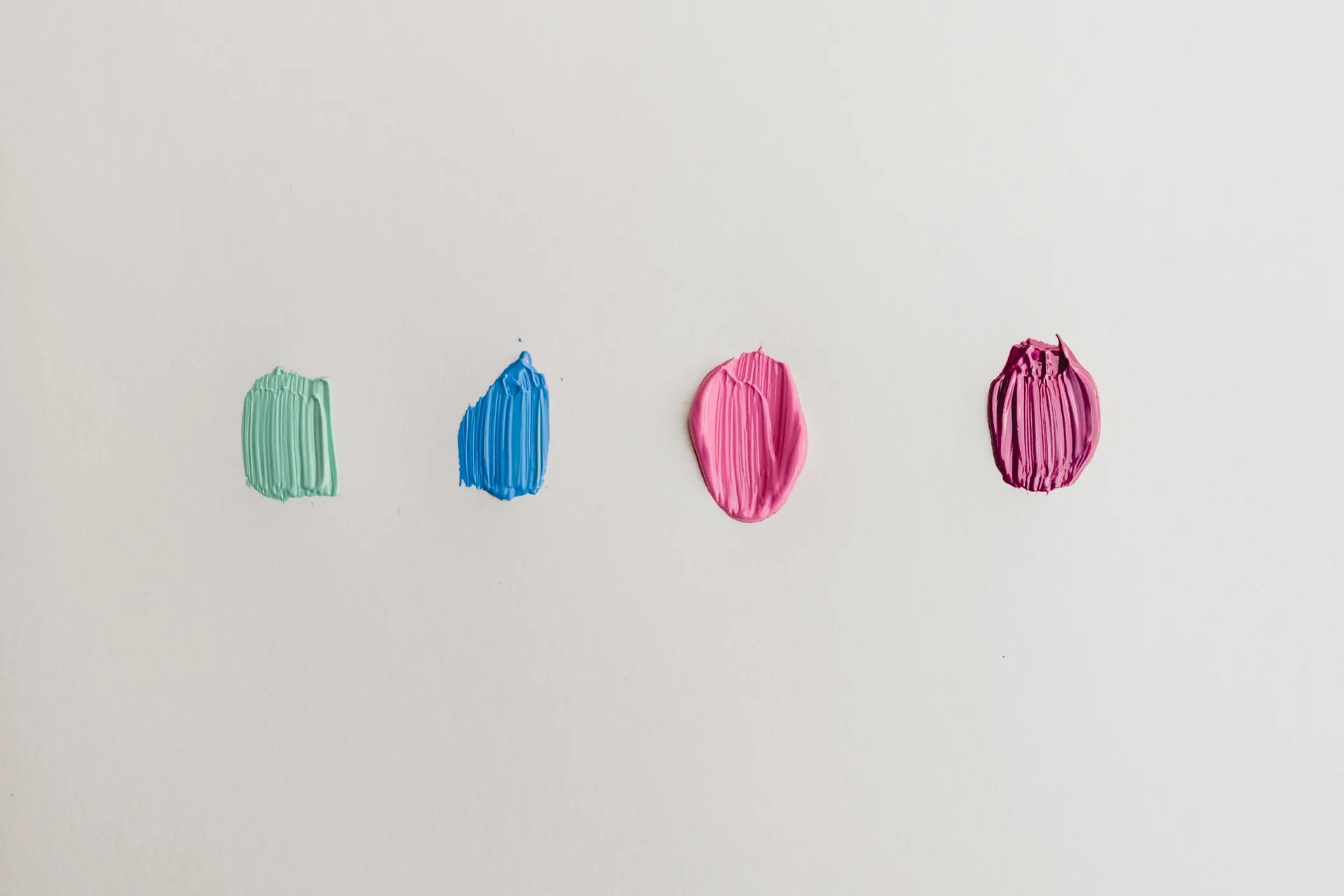 Four acrylic paint smears on white sheet in mint green blue pink and magenta with visible brush stroke texture spaced in a row