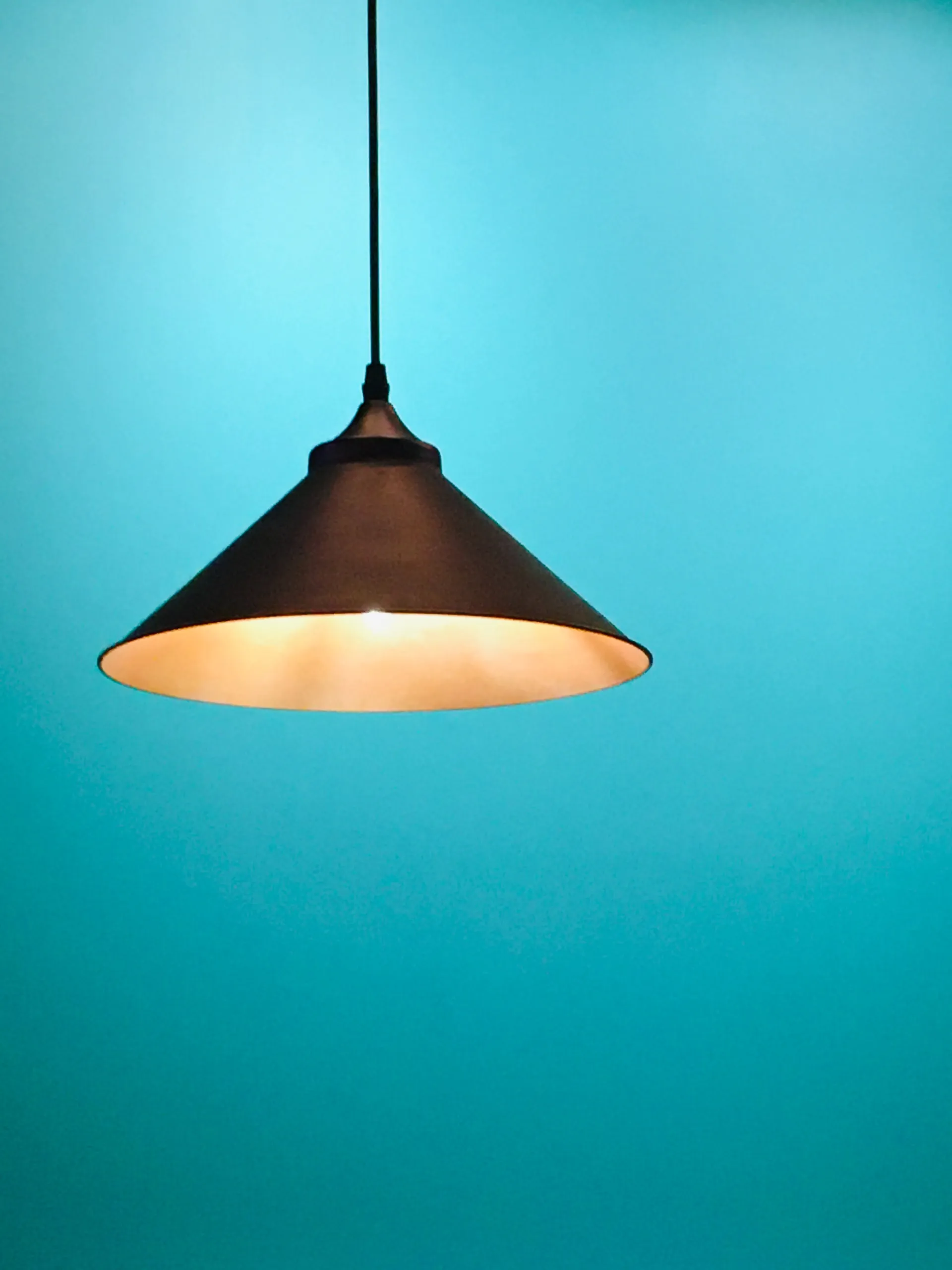 Minimalist black metal cone pendant light fixture glowing softly against a smooth turquoise wall