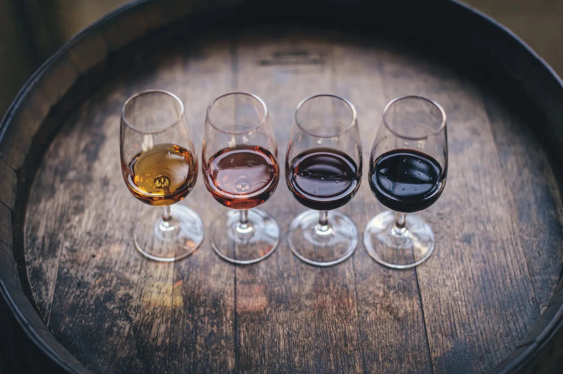 Four wine glasses filled with orange, rosé, light red and dark red wine arranged in a row on a rustic wooden barrel surface