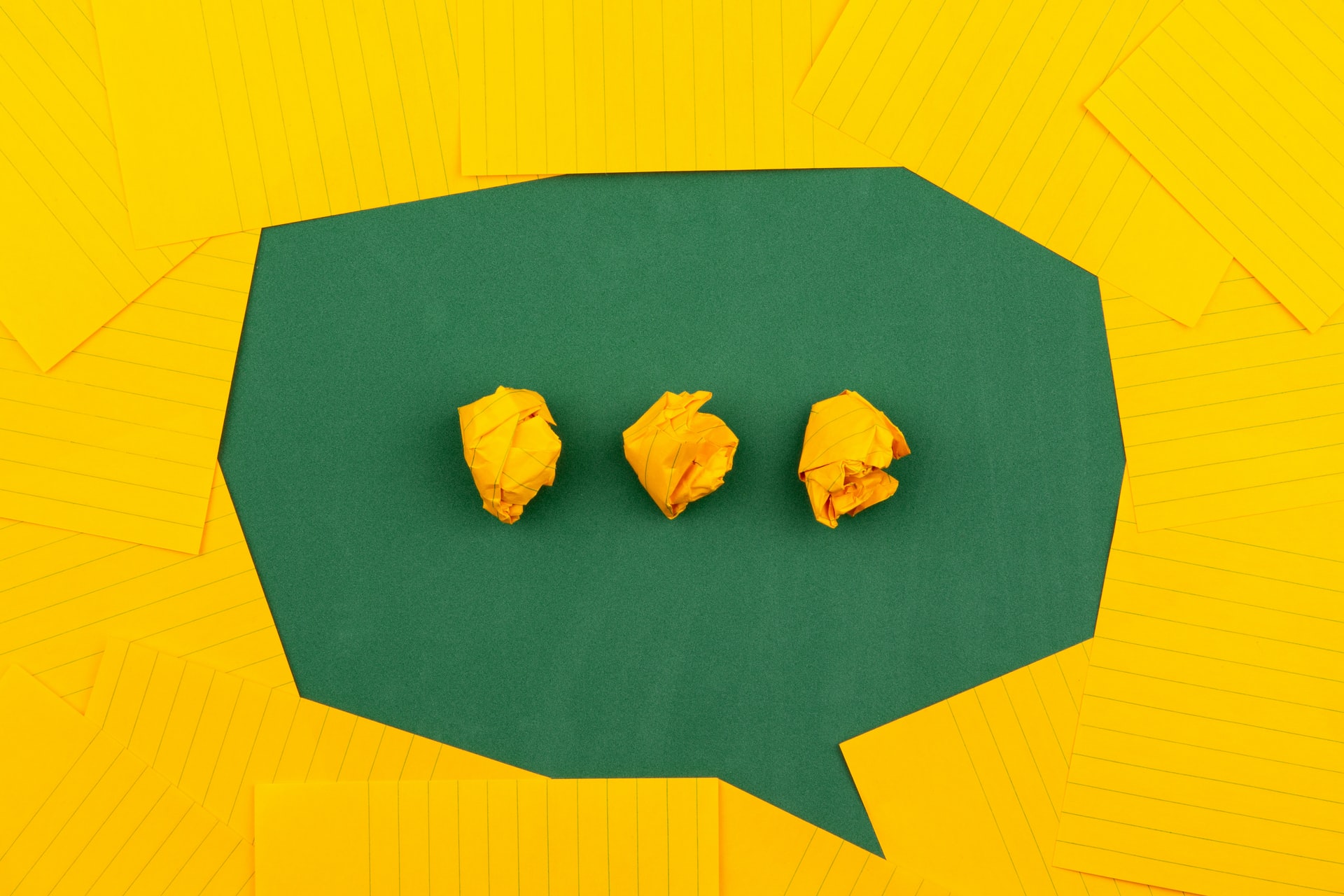 Overhead view of a green paper speech bubble shape framed by scattered yellow lined sheets with three crumpled yellow paper balls aligned as a typing ellipsis