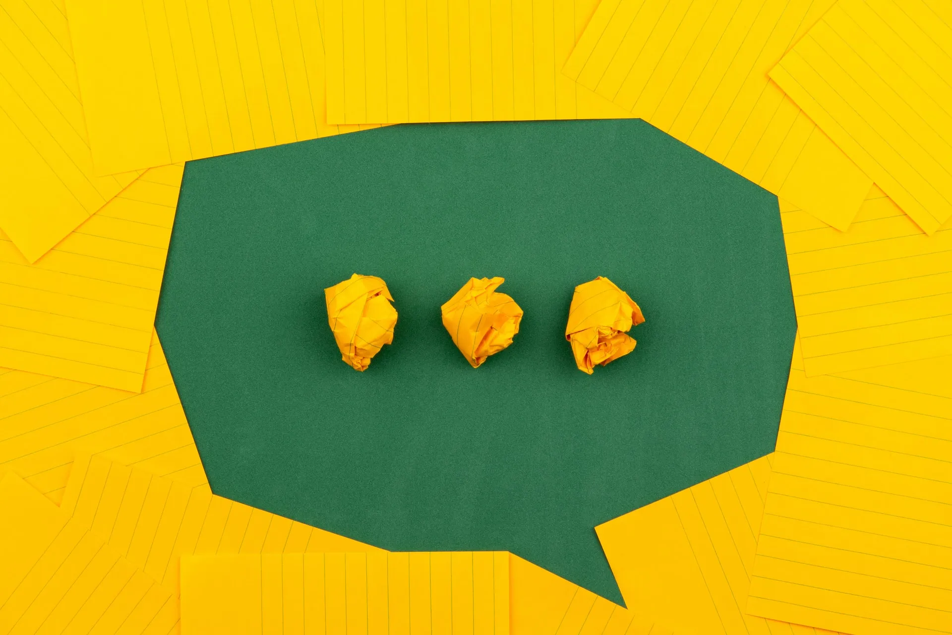 Overhead view of a green paper speech bubble shape framed by scattered yellow lined sheets with three crumpled yellow paper balls aligned as a typing ellipsis