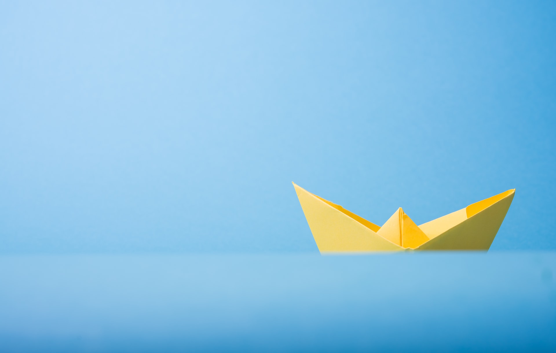 Yellow paper boat floating on a smooth blue surface against a light blue backdrop