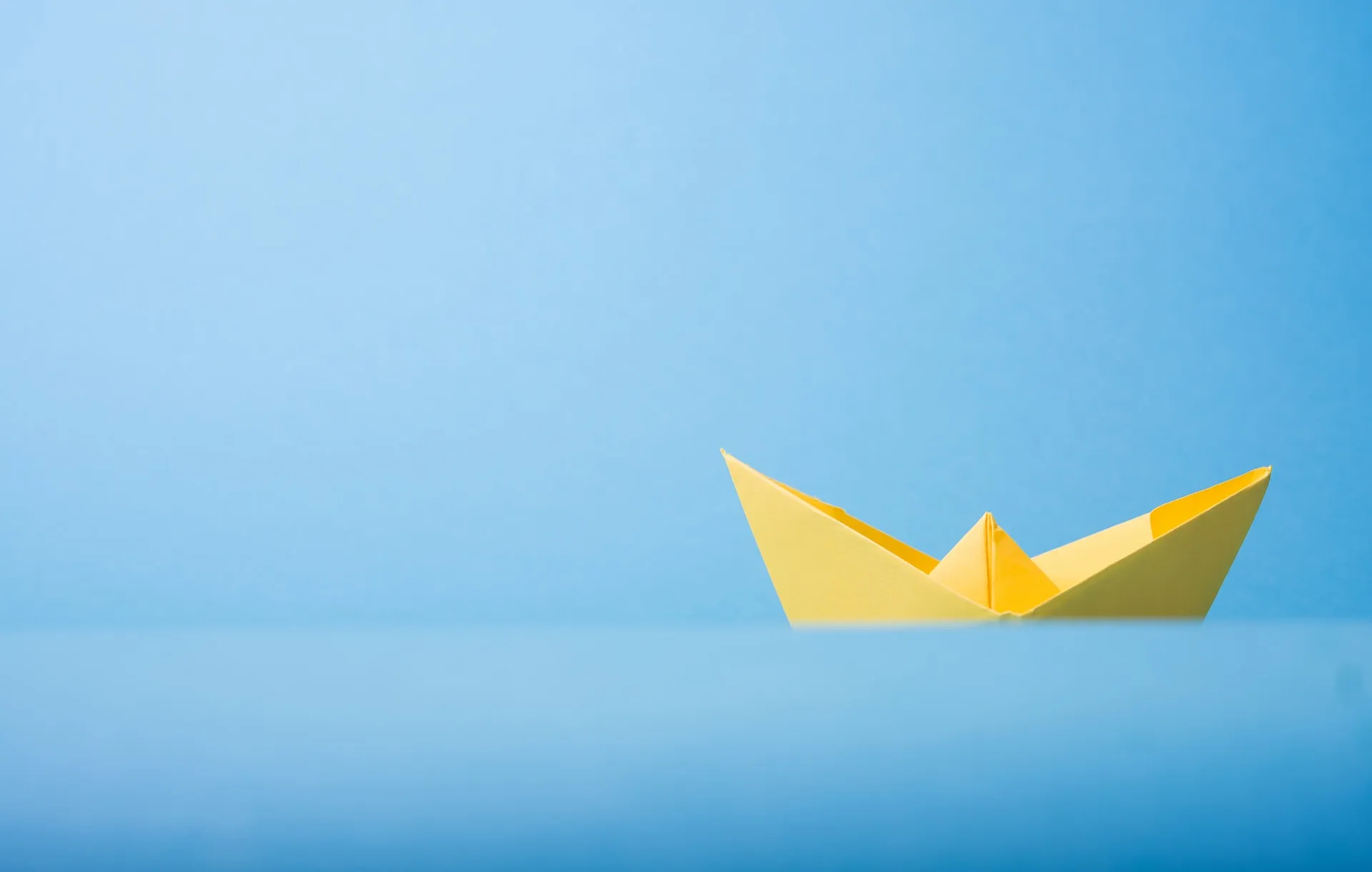 Yellow paper boat floating on a smooth blue surface against a light blue backdrop