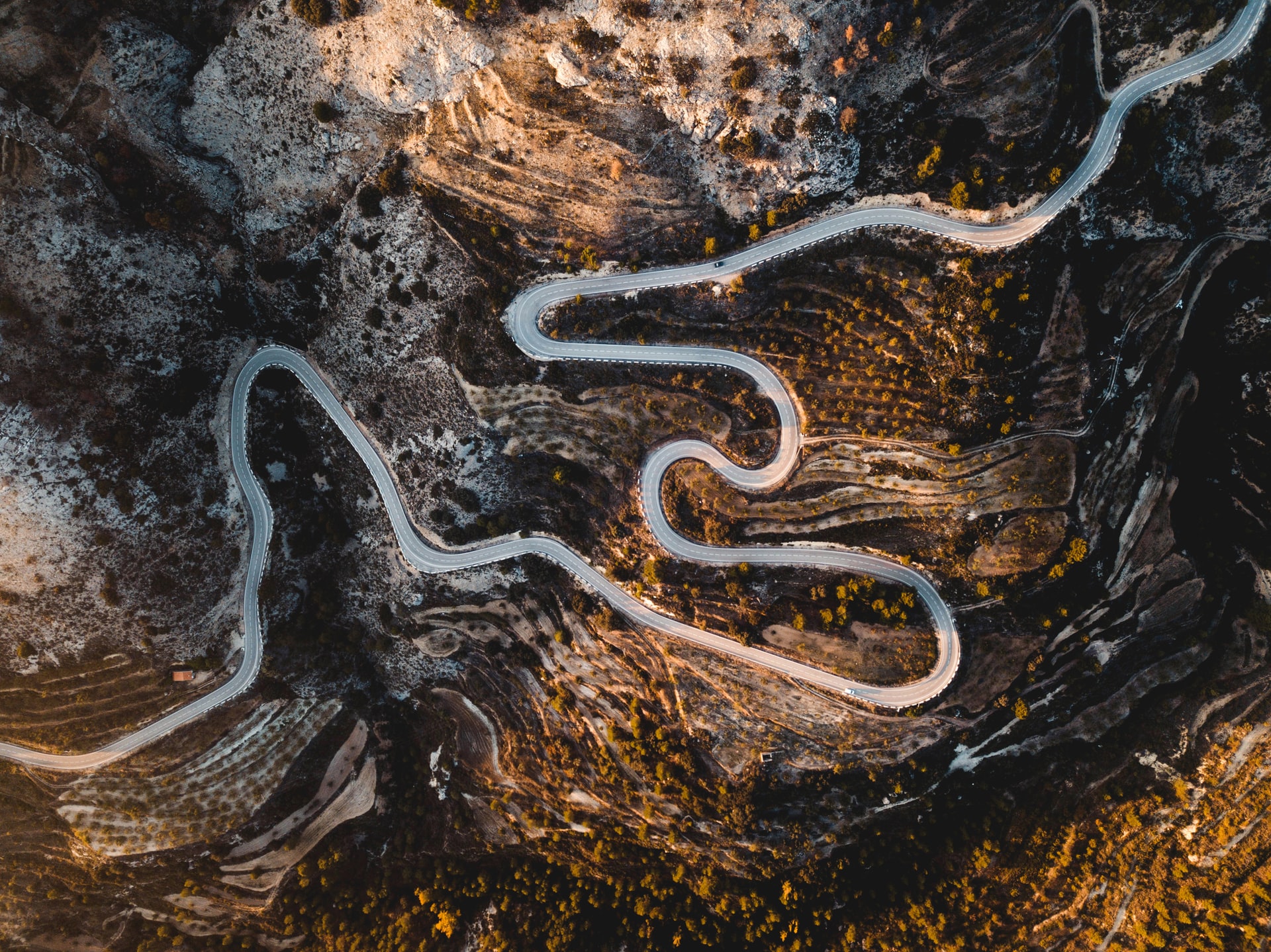 High aerial view of a serpentine mountain road snaking through rugged rocky terrain with sparse vegetation under warm light