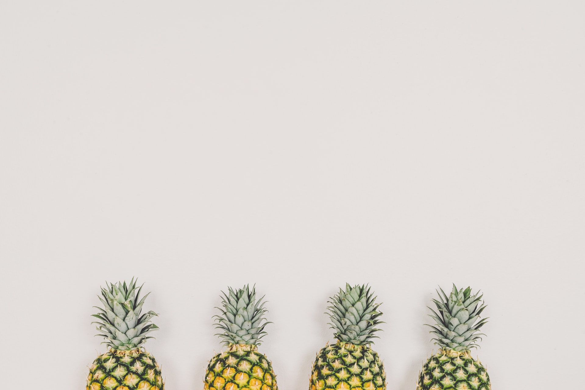 Four ripe pineapples arranged side by side along the lower edge of a pale cream backdrop