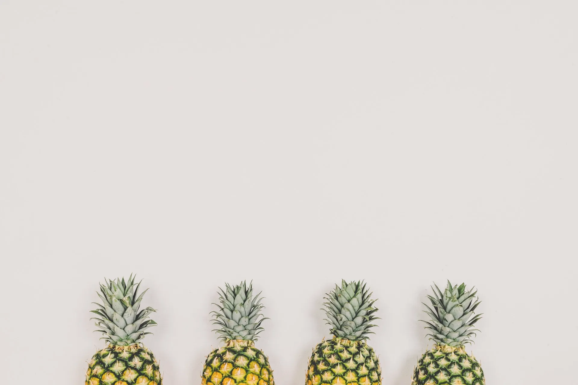 Four ripe pineapples arranged side by side along the lower edge of a pale cream backdrop