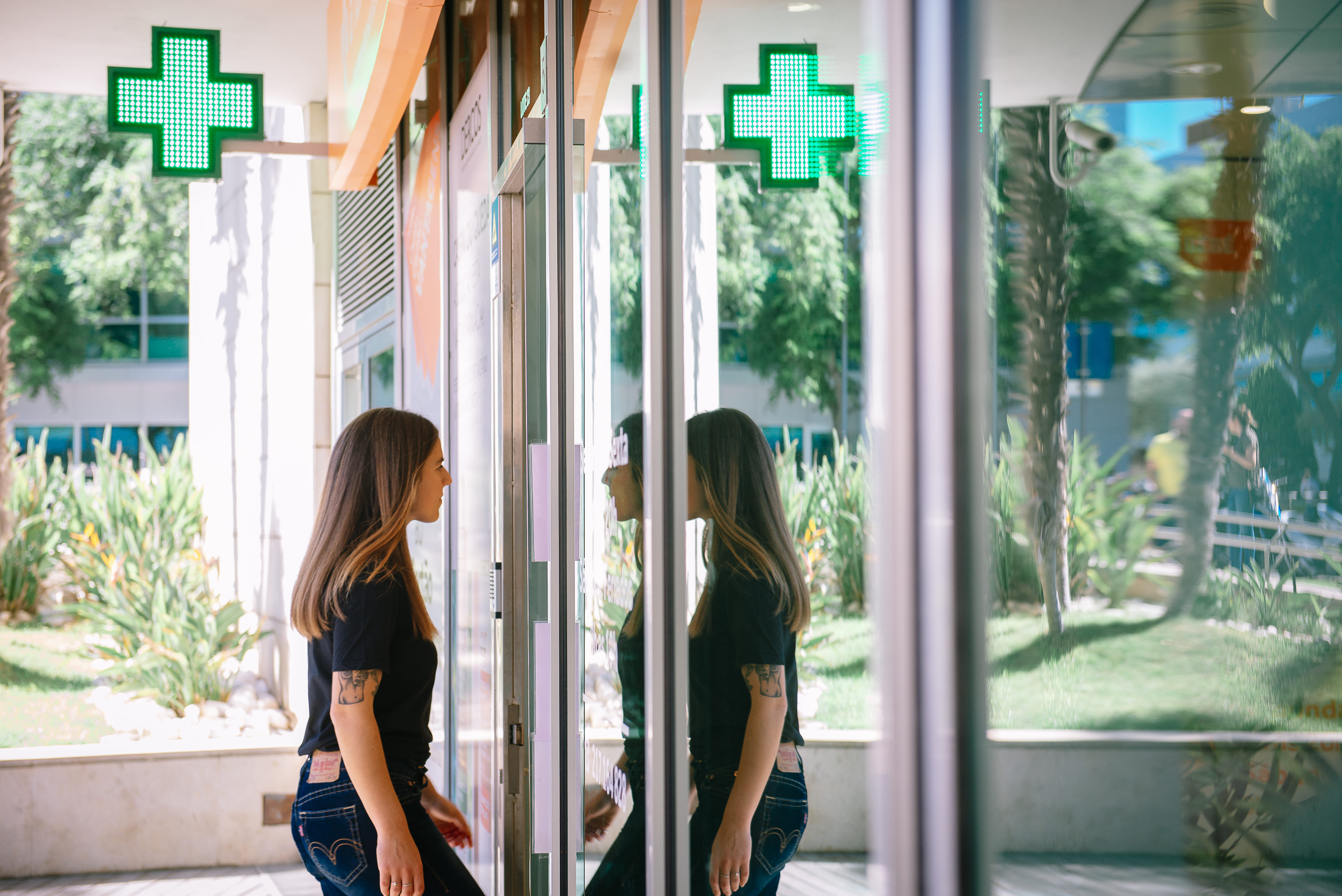 Mujer de perfil con tatuajes camiseta negra apoyada en la puerta de cristal de una farmacia iluminada con cruces verdes con su reflejo en el espejo y fondo ajardinado urbano