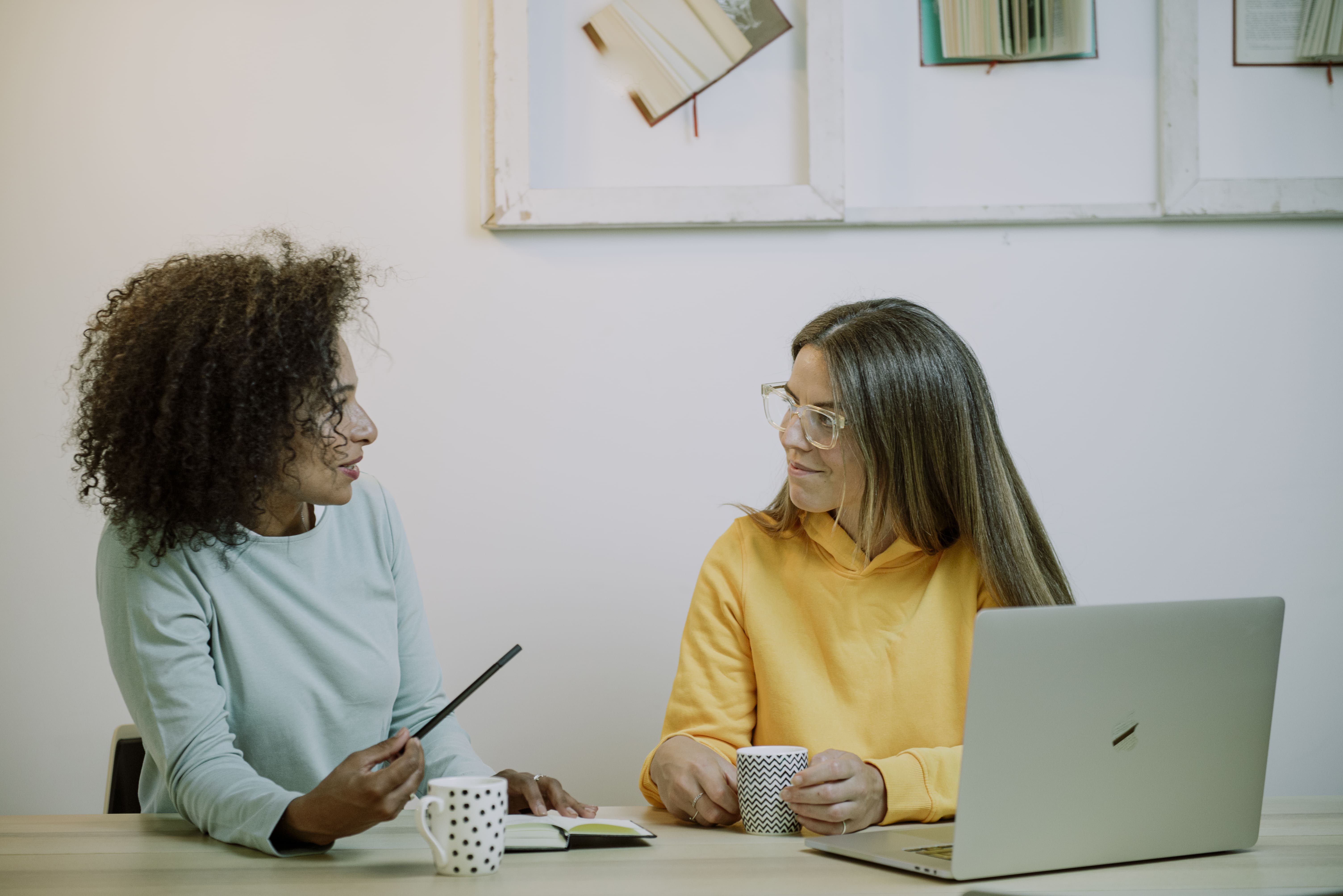 Dos compañeras de trabajo conversando en una mesa con tazas de café libreta y ordenador portátil frente a libros abiertos colgados en la pared de fondo