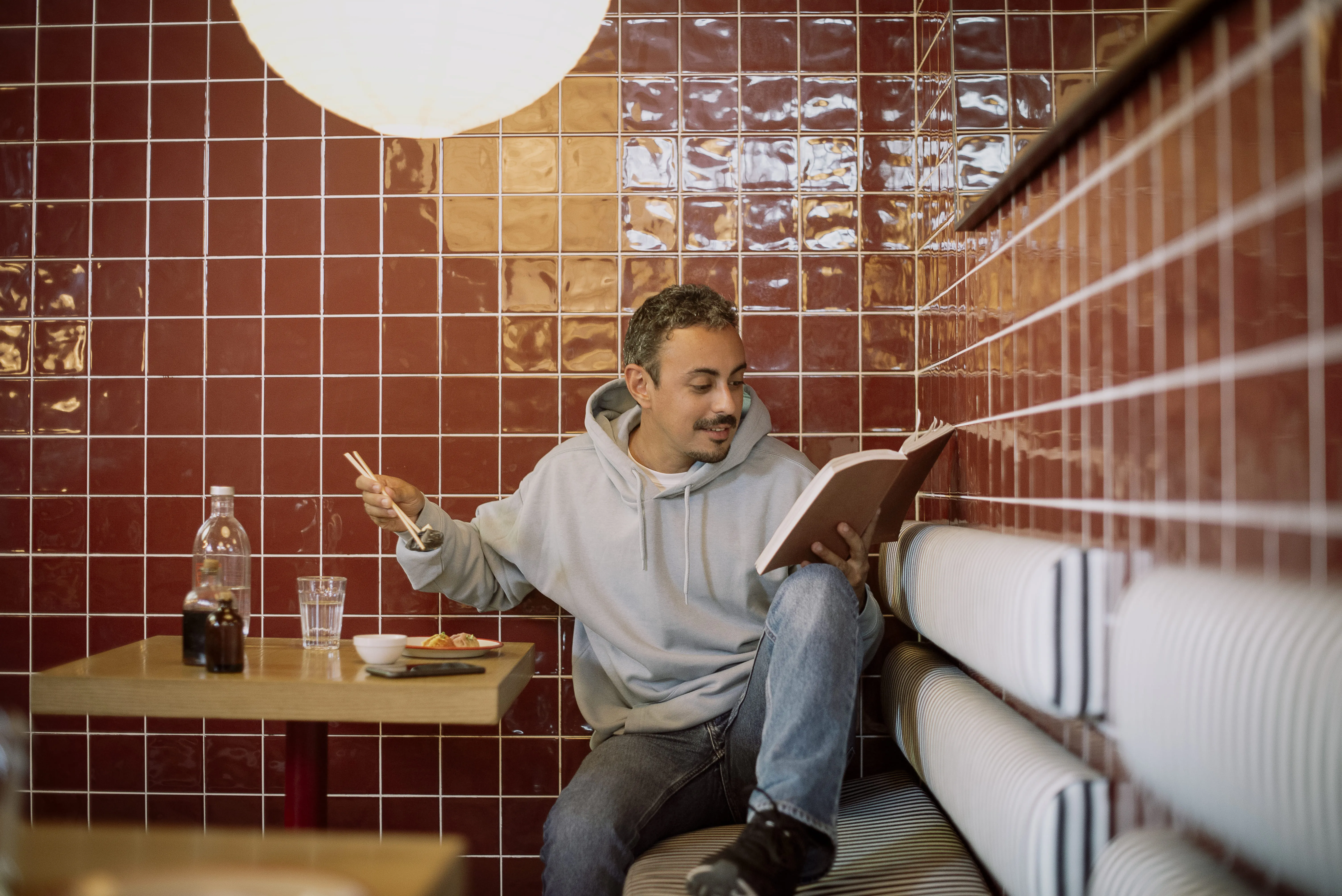 Hombre sentado con plato de sushi y libro en restaurante de azulejos rojos