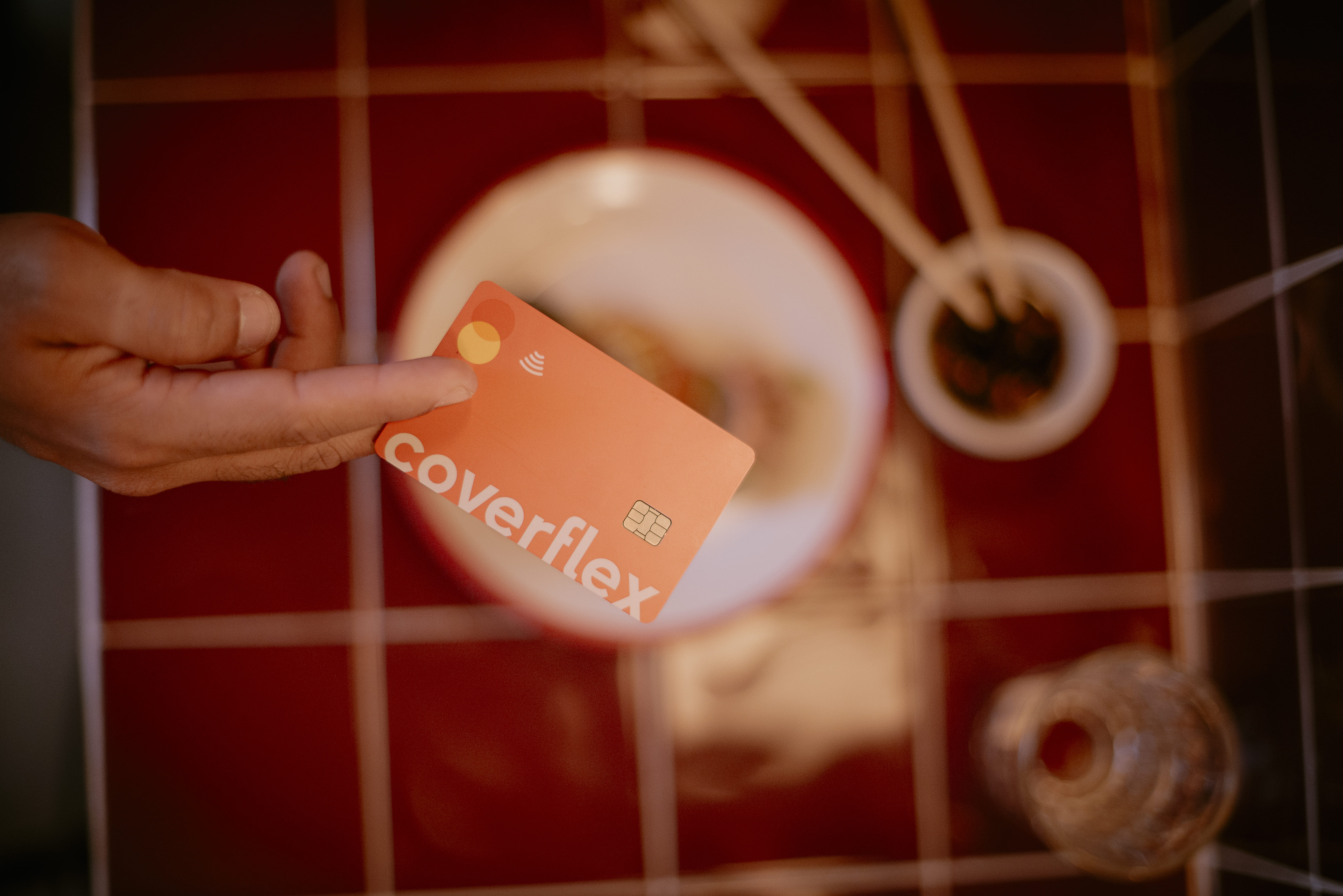 mano sosteniendo tarjeta Coverflex naranja con logotipo chip y simbolo contactless sobre mesa de azulejos rojos junto a un cuenco de comida asiatica con palillos