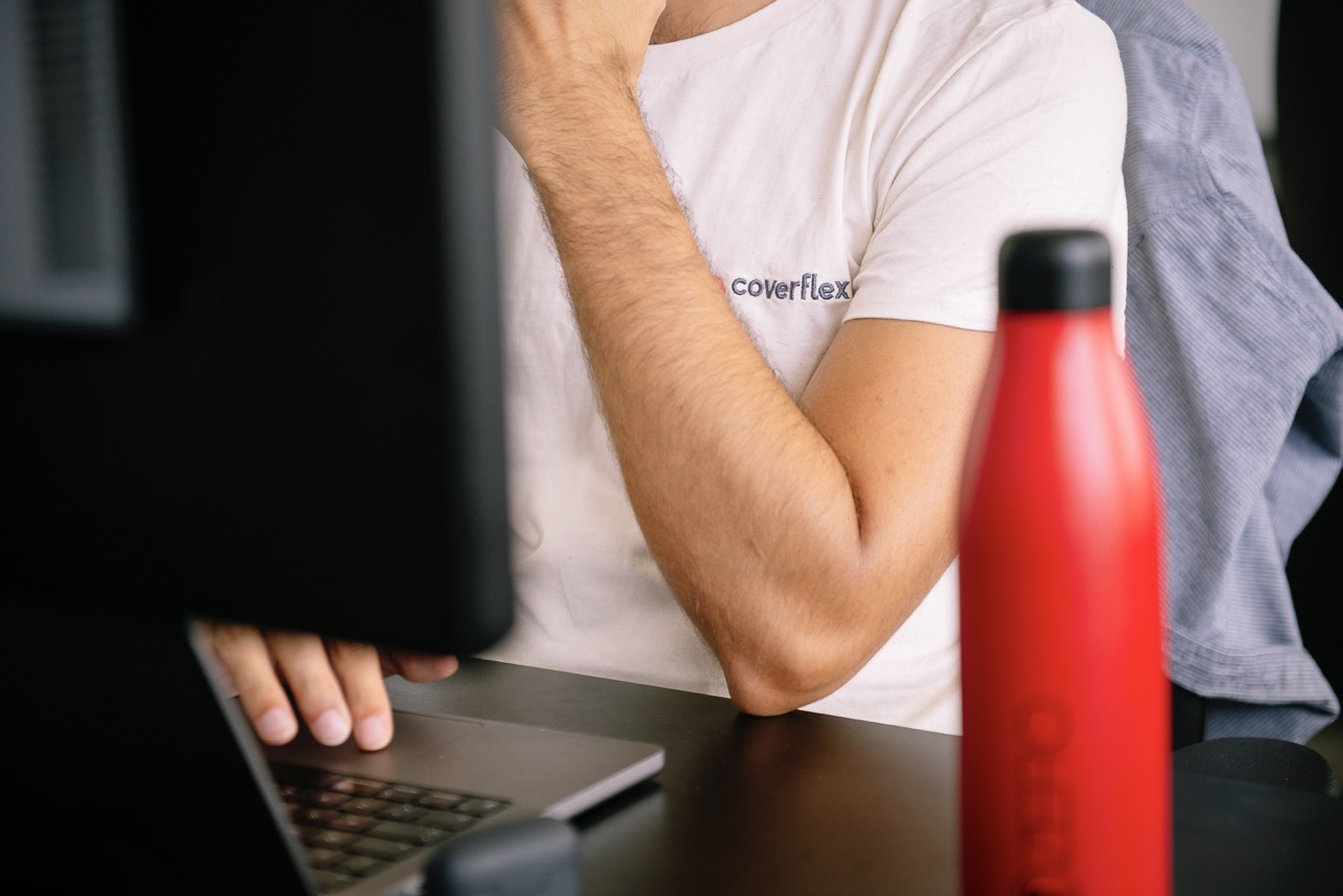 Vista parcial de un empleado con camiseta Coverflex trabajando en un portatil con monitor delante y botella termica roja sobre el escritorio