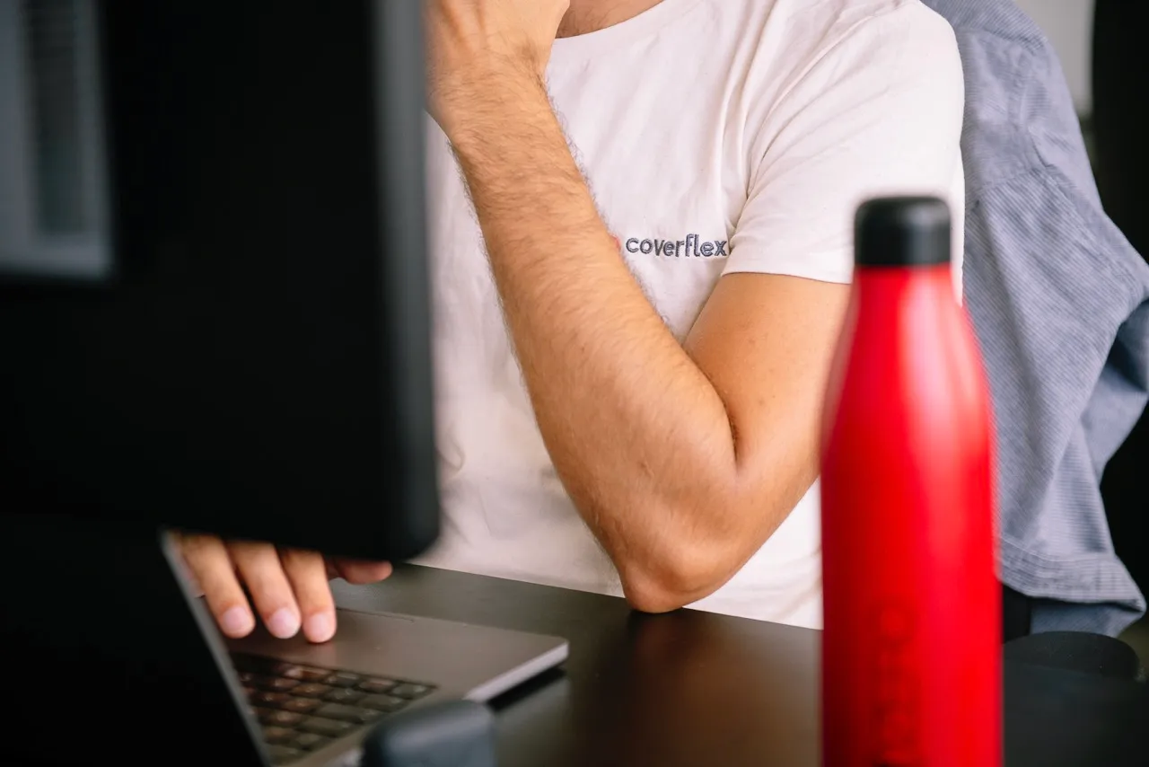 Vista parcial de un empleado con camiseta Coverflex trabajando en un portatil con monitor delante y botella termica roja sobre el escritorio