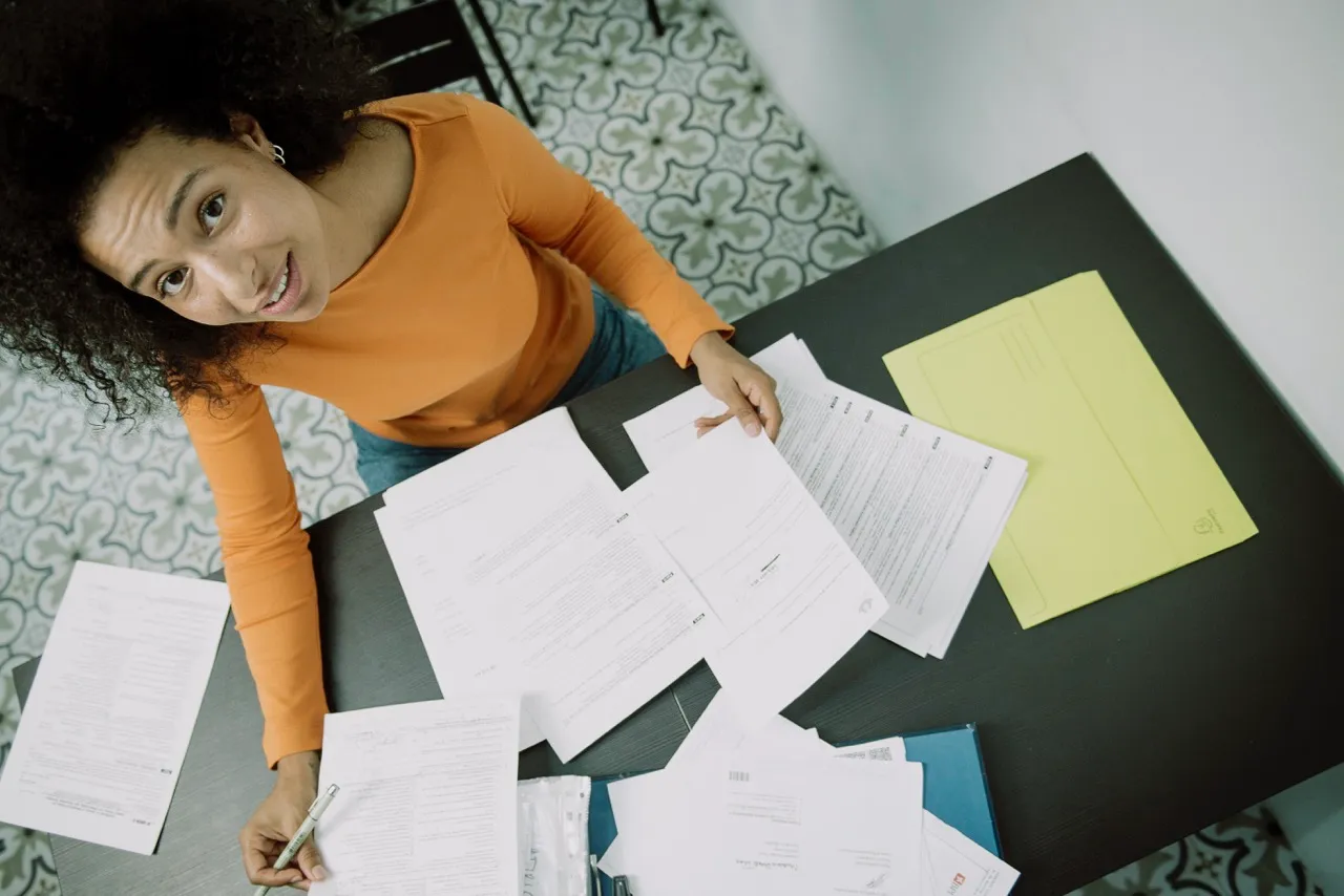Mujer sentada en un escritorio con varios documentos y carpetas, organizando formularios mientras mira hacia la cámara