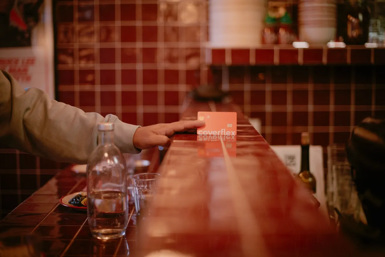 mano de persona apoyada en barra de bar junto a tarjeta naranja Coverflex en foco con azulejos rojos y utensilios desenfocados