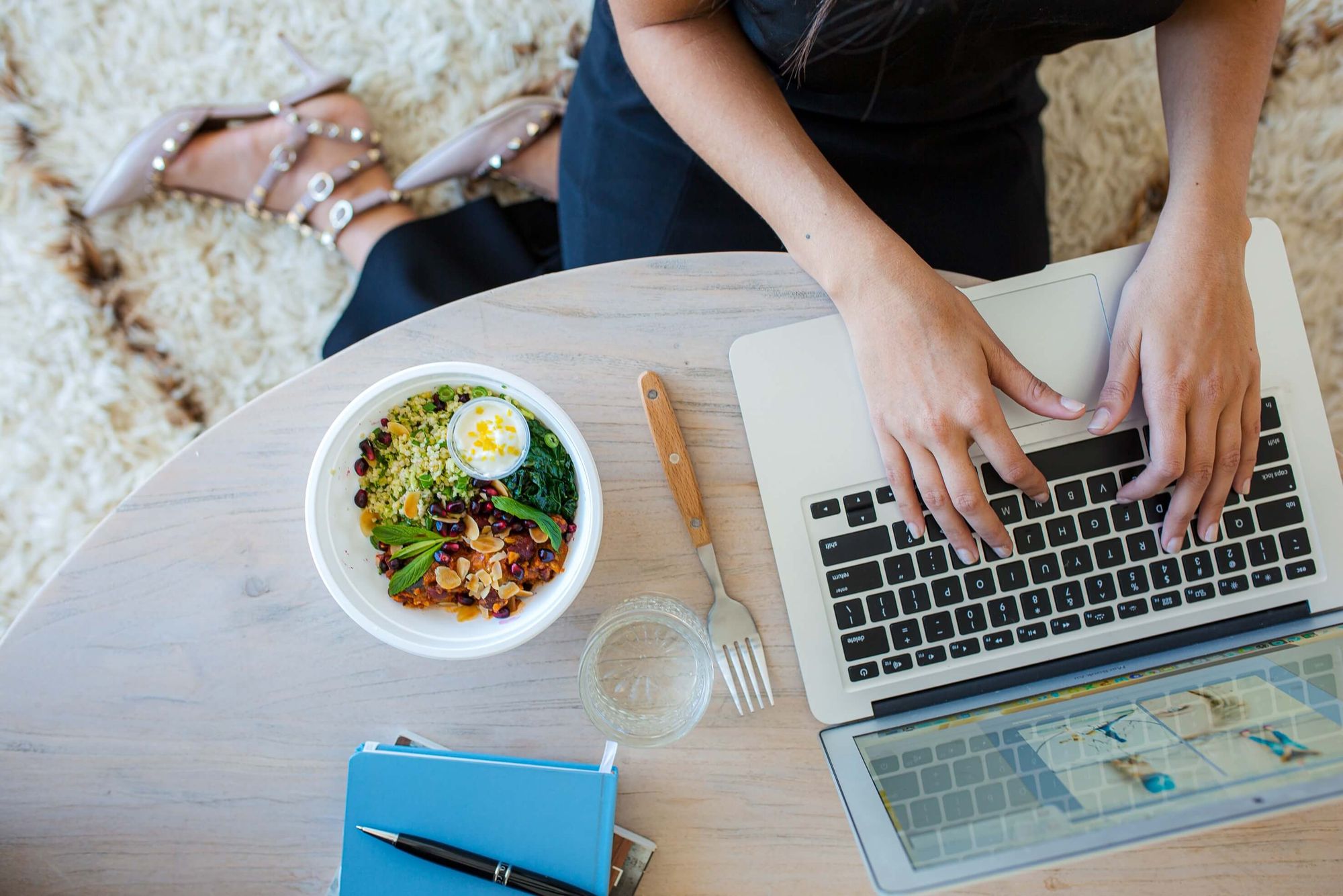 Vista cenital de manos femeninas escribiendo en un portatil sobre mesa de madera junto a un tazon con ensalada colorida un tenedor un vaso de agua y un cuaderno azul